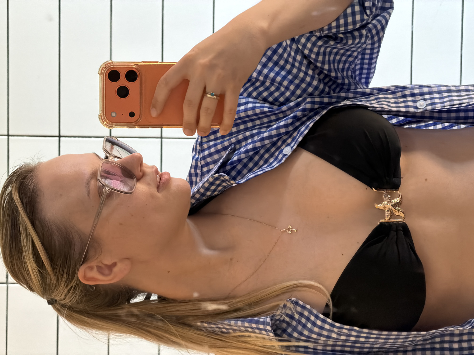 A fair-skinned woman takes a selfie in front of a white-tiled wall. She wears glasses, a black bikini top with a gold starfish ornament, and a blue and white checkered shirt. The phone case is orange. A gold ring is on her finger. A gold necklace with a key pendant adorns her neck. The location cannot be determined from the image.