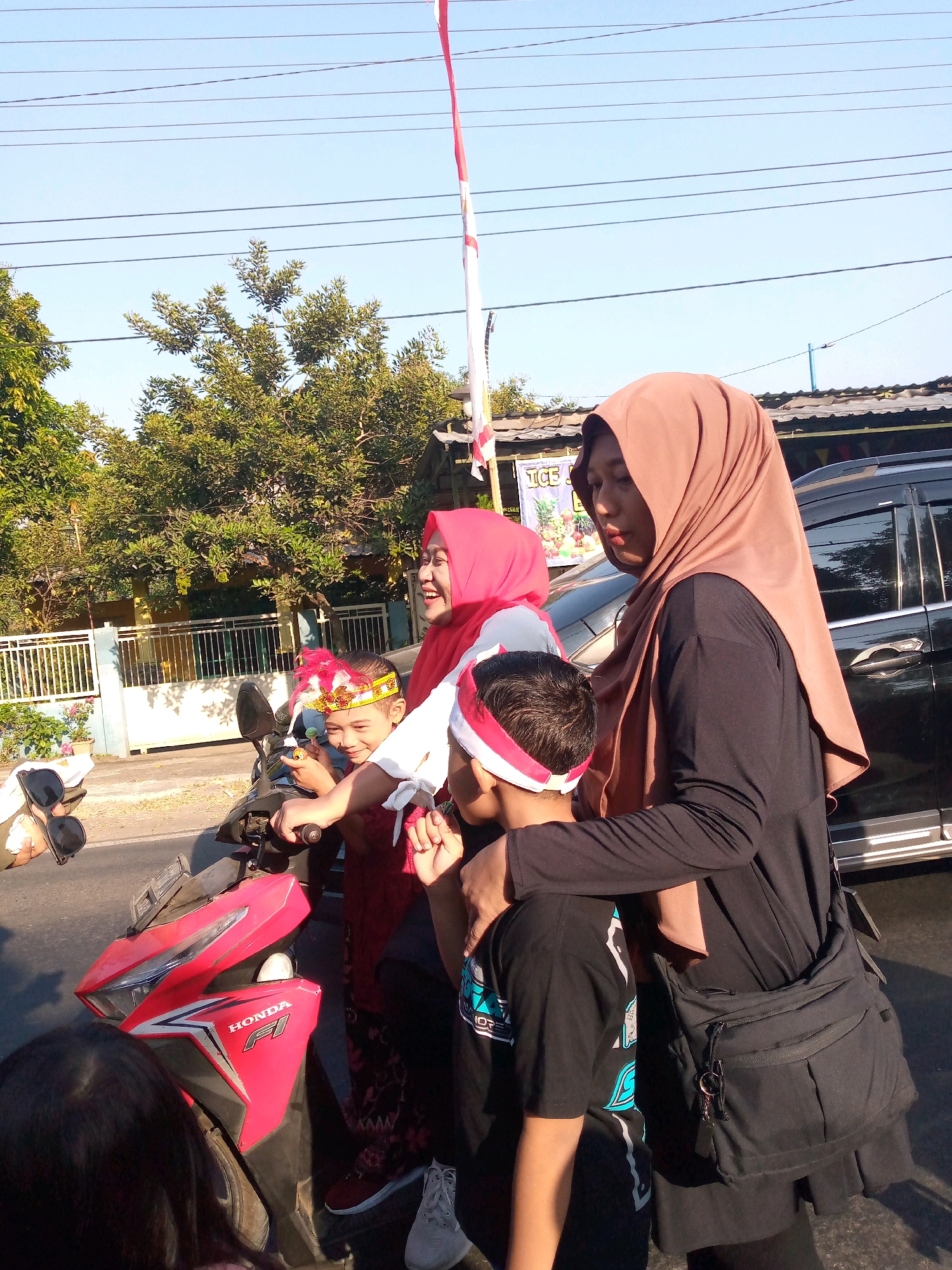 In Sumberwono, Indonesia, under a clear blue sky with power lines overhead, a group of individuals, likely a family, gathers near a red Honda F1 motorcycle. Two women, one wearing a pink hijab and the other a brown hijab, are present. The woman in the brown hijab has her arm around a young boy, who is wearing a red and white headband and a black t-shirt with blue lettering. He is seated on the motorcycle and appears to be holding something in his right hand. Another child, possibly a girl, with a red and gold headband, is also visible on the motorcycle, looking towards the viewer. A red and white Indonesian flag flies from a pole in the background, suggesting a celebration or national event. A black SUV is parked to the right of the group. The environment suggests a street scene, possibly a residential area or a roadside gathering.