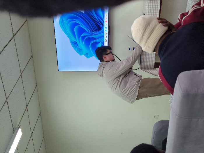 A medium shot captures a person in a light tan jacket and glasses standing and reaching toward a large monitor displaying a blue design. Part of the monitor is obscured by a blurry object at the top of the frame. To the right, a person wearing a white beanie, a dark blue shirt, and a red-striped sleeve is partially visible. The setting appears to be indoors, with a gray chair visible in the foreground and a tiled ceiling and a long fluorescent light on the left side of the shot.