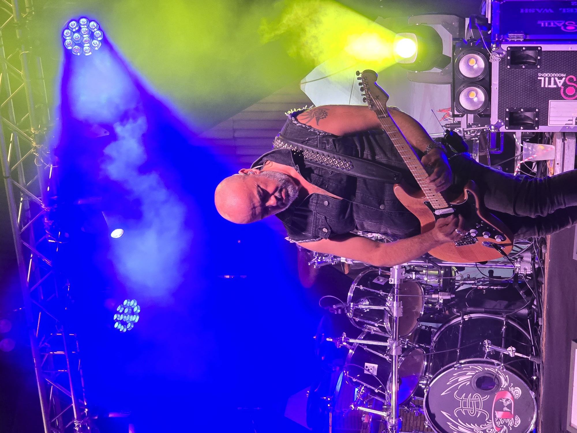 The image depicts a rock concert at a venue in Cornellà de Llobregat, Spain, possibly the Parc de Can Mercader, given the presence of concert lighting and a stage setup. A bald, middle-aged man with a goatee is playing an electric guitar on stage. He is wearing a black denim vest over a black t-shirt, black jeans, and a studded guitar strap. His left arm, visible with a tattoo, is positioned to play the guitar. Behind him, a drum kit with various cymbals and drums is set up. The stage is illuminated by bright blue and green stage lights, with smoke or fog effects contributing to the atmosphere. In the foreground, to the right of the guitarist, are speaker cabinets and lighting equipment, including two prominent white lights on one of the speakers. A cup with a straw and a drink is visible on a white surface to the left of the speaker setup. The overall impression is that of a dynamic live music performance.