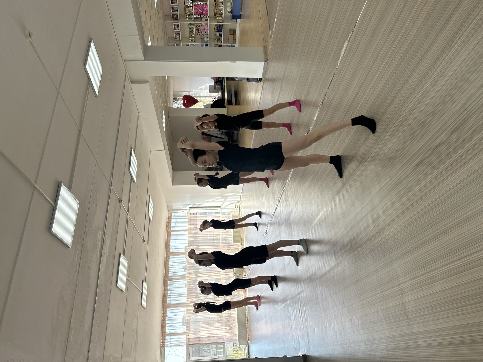In Votkinsk, Russia, a group of seven individuals dressed in black shirts and shorts practice a dance or exercise routine in a studio with a light-colored, lined floor. They are positioned in a row, facing the same direction, with their arms raised above their heads. Some wear pink socks while others wear black or white. The room is brightly lit by a series of square ceiling lights. Windows line the left side of the room. In the background, there's a shelf with various items and a red heart-shaped balloon. The scene appears to be a dance or fitness class in progress.