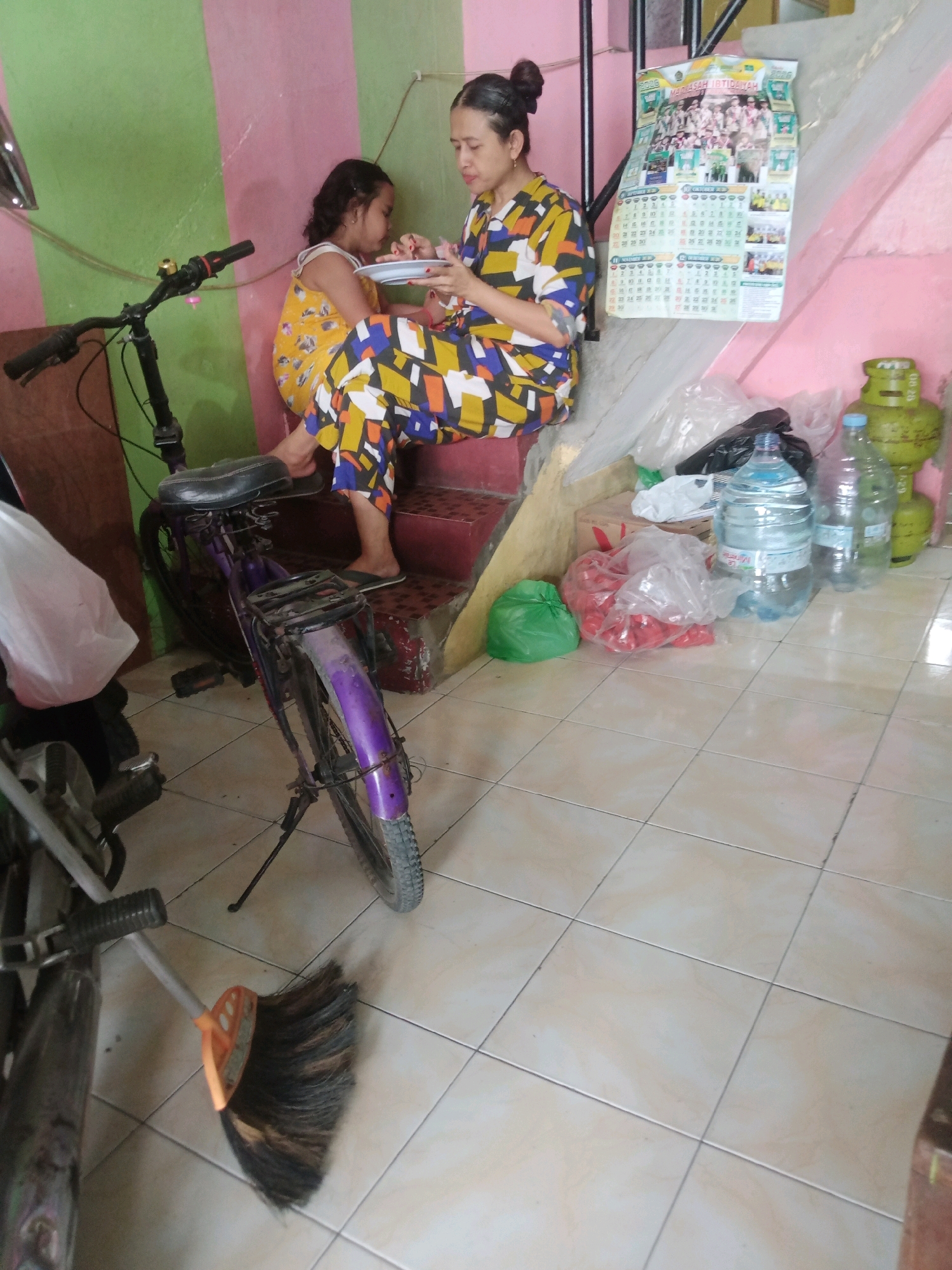 In a room with a green and pink striped wall, a woman in a colorful patterned outfit sits on a step and feeds a young girl. The girl, wearing a yellow dress, sits beside her. In the foreground, a purple bicycle is parked. Various items are scattered around, including large water bottles, plastic bags filled with red items, a green plastic bag, a gas cylinder, and a broom. A calendar hangs on the wall behind the woman. The floor is tiled.