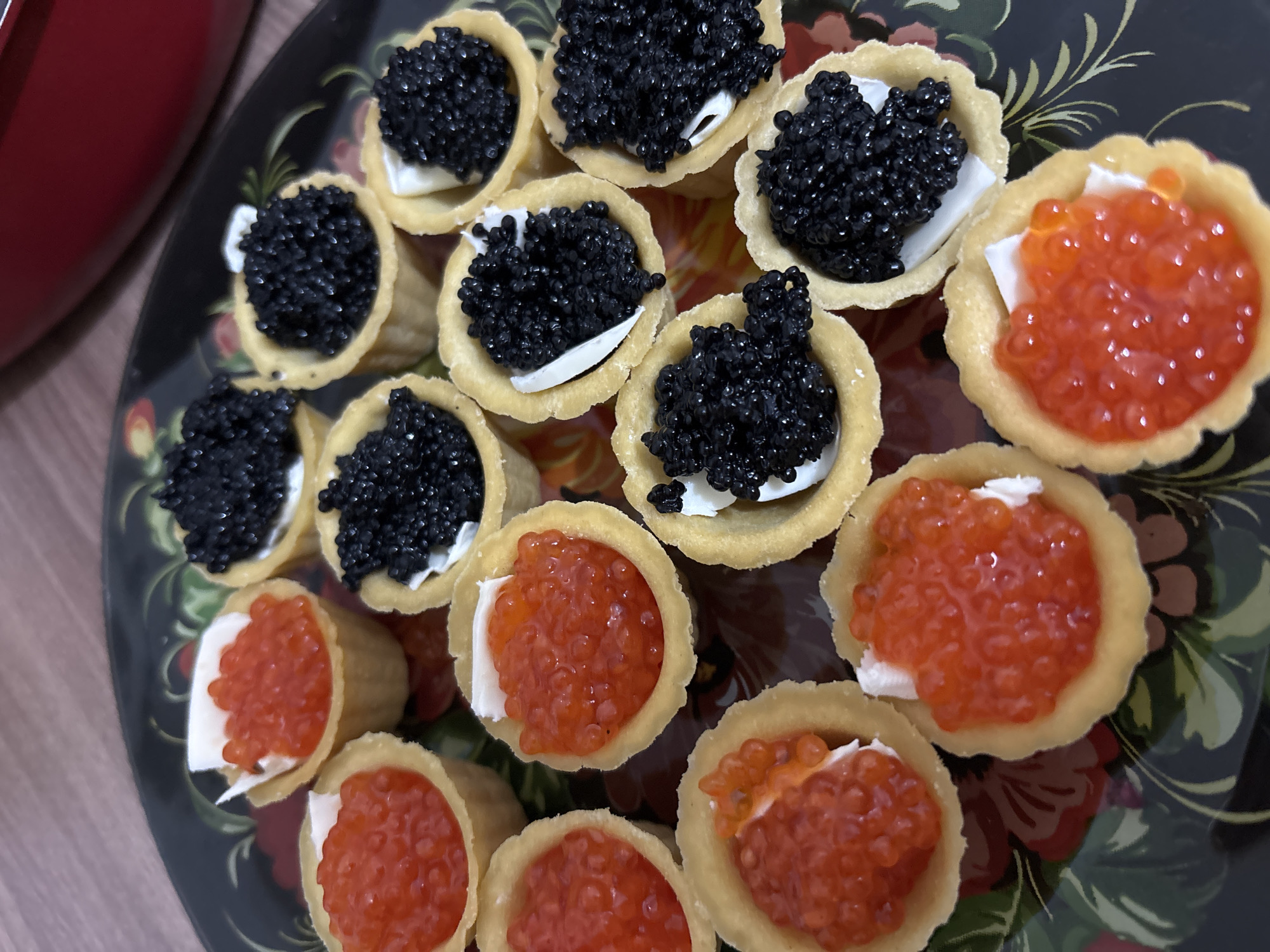 A high-angle, close-up shot shows a platter of canapés topped with black and red caviar. These are served in small, golden pastry shells. A base of white cream cheese is visible under both types of caviar. The platter has a decorative floral pattern painted on its surface. The shot is taken from above and at an angle, with a section of a dark red object appearing in the top left corner. The background is a light-colored wooden surface. This food is likely being presented in Sochi, Russia.