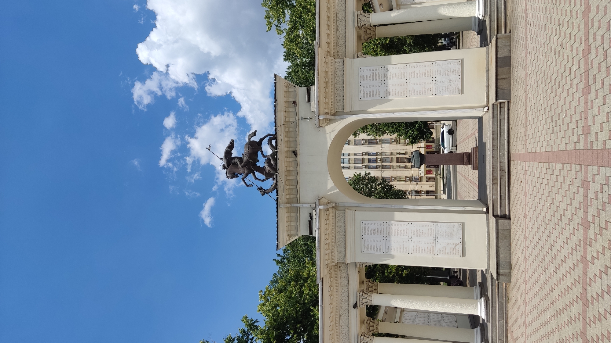 The image shows an outdoor view of the Monument to the Liberators of Kuban in Krasnodar, Russia. The monument features a large archway with Corinthian columns, and atop the arch is a bronze sculpture of St. George slaying a dragon. On the walls within the archway are inscribed lists of names. In the background, through the arch, a modern building with many windows is visible, along with trees and a white car. The ground in front of the monument is paved with interlocking bricks. The sky is blue with scattered white clouds.