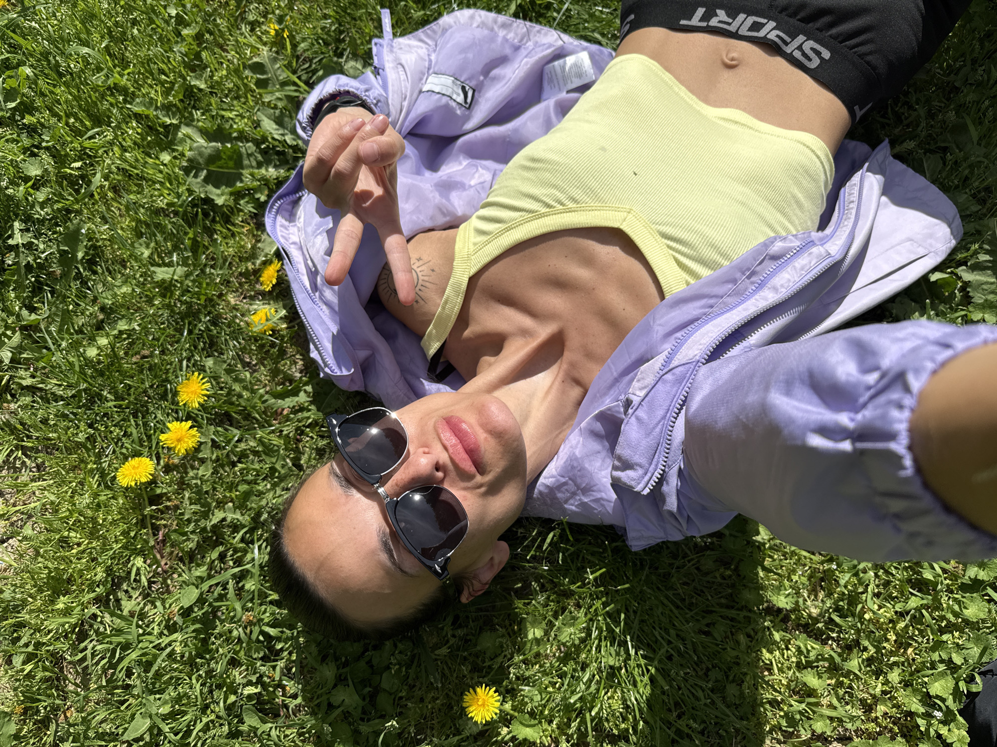 An overhead, slightly tilted shot captures a person lying on their back in vibrant green grass under bright sunlight in Pleven, Bulgaria. The individual is positioned diagonally across the frame, looking up towards the camera. They wear dark sunglasses pushed up on their nose, covering their eyes, and their lips are slightly parted.
The person is dressed in a light yellow ribbed crop top and a black sports bra, which has the word "SPORT" visible upside down across the band. Black shorts or underwear are also visible. A light purple, possibly windbreaker-style jacket is draped around their upper body, with one sleeve partially off. A small, stylized sun and moon tattoo is visible on their left upper arm. Their left hand is raised, forming a V-sign with two fingers.
The setting is a lush grassy area, scattered with several bright yellow dandelions. The strong illumination and distinct shadows suggest a sunny day, likely during the mid-day or afternoon. The overall impression is one of relaxation and enjoying the outdoor environment. A clothing label can be faintly seen inside the purple jacket.
