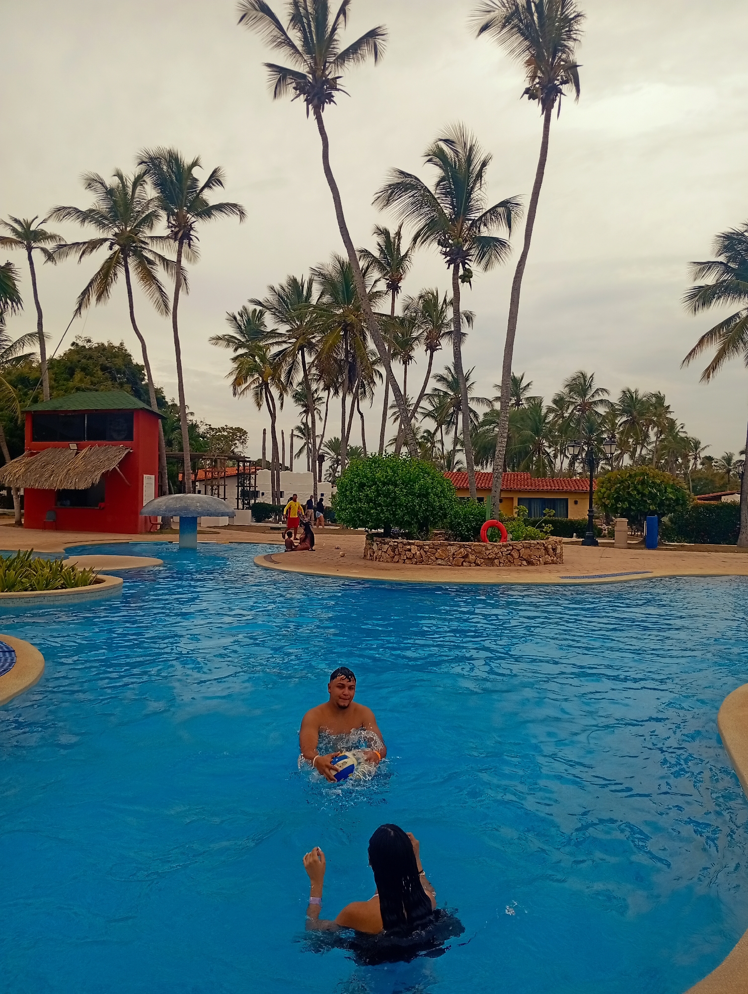 The image depicts a swimming pool in Playa El Agua, Venezuela. Two individuals are in the pool: one male with a ball and a female facing away from the camera. The pool is surrounded by palm trees, structures, and landscaping. In the background, there is a red building with a thatched roof section. Other buildings and people can be seen further back. The sky is overcast.