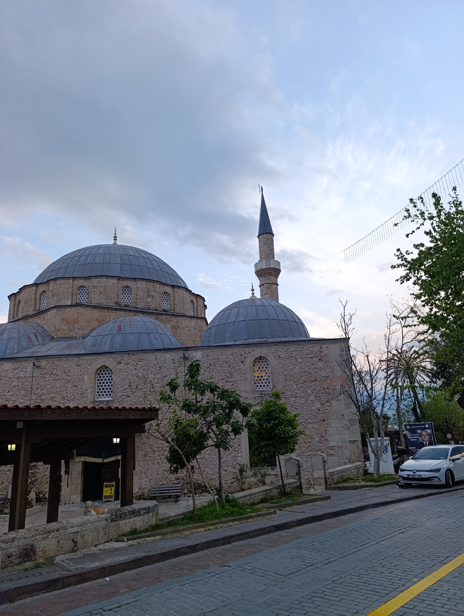 The image depicts a mosque with a minaret and multiple domes in Antalya, Turkey. The mosque is constructed of light-colored stone. A white car is parked on the cobblestone street in front of the mosque. The sky is overcast. A covered structure is attached to the mosque. Trees are present on the side of the road. A poster stands near the car.