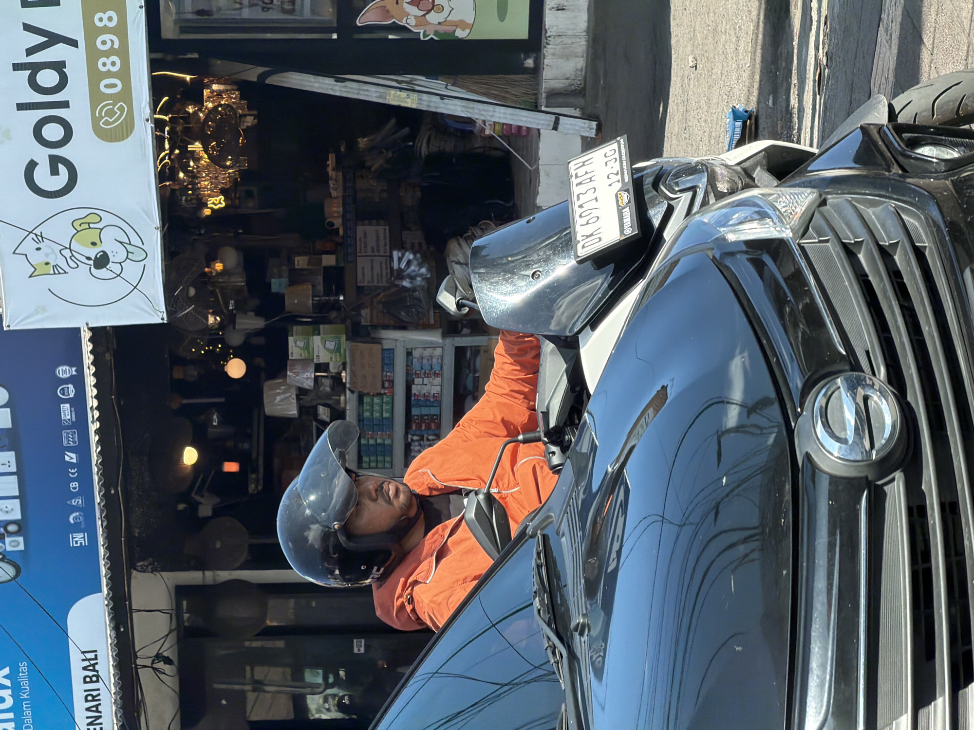 An individual, likely an adult male, wearing a black open-face helmet with a clear visor and a bright orange jacket, is seated on a scooter. The scooter is partially visible, appearing to be black and possibly silver, with a license plate reading "DK 6913 AFH" and an expiry date "12-30". A "YAMAHA" logo and the slogan "SEMAKIN DIDEPAN" (meaning "further ahead") are visible on the license plate holder.
The scooter is positioned next to a black car, the front hood, windshield, and a portion of the grill of which are visible in the foreground. Reflections of power lines and a bright blue sky are prominent on the car's glossy surface, indicating clear and sunny weather.
In the background, there are storefronts lining a street in North Kuta, Indonesia. One sign, predominantly white, displays "Goldy" and a phone number "0898," along with illustrations of a cat and a dog, suggesting a pet-related business. Another partial blue sign shows text like "Dalam Kualitas" and "ENARI BALI," accompanied by various certification logos including CB, CE, SNI, and "LED." Inside the shops, various merchandise, including lighting fixtures, can be seen, with a ladder leaning against a wall in one of the establishments. The scene depicts a typical busy street environment.
