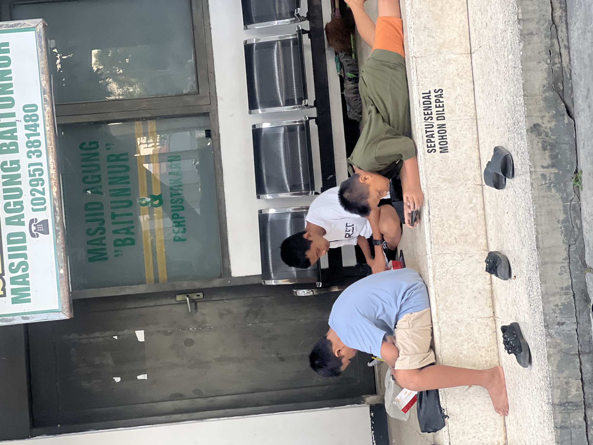 This image shows three young males in front of a building with a sign that reads "MASJID AGUNG BAITUNNUR" and a phone number, likely in Kecamatan Pati, Indonesia. The building appears to be a mosque and library, indicated by the text "MASJID AGUNG BAITUNNUR & PERPUSTAKAAN" visible through a window. Two of the individuals are seated on steps, while the third is lying prone on the ground. The two seated individuals are engaged with their mobile phones. The prone individual is also looking at a mobile phone. In front of the individuals, there are various footwear items scattered on the steps, including black sneakers and black sandals. A sign on the steps reads "SEPATU/SENDAL MOHON DILEPAS," which translates to "Shoes/Sandals Please Remove." The environment suggests an outdoor waiting or resting area.