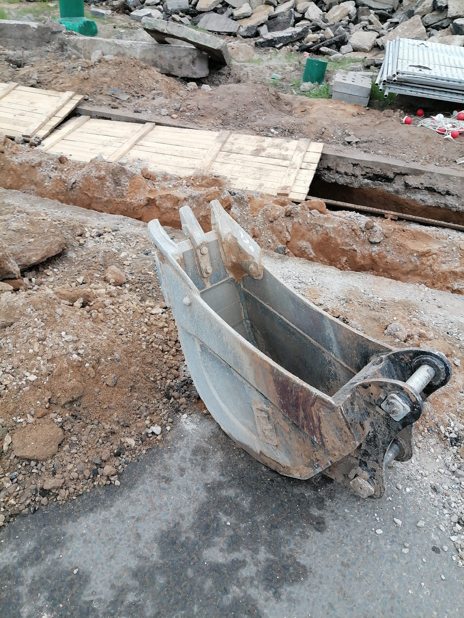 The image shows an excavator bucket resting on an asphalt surface at a construction site. To the left of the bucket, there is a mound of dirt and rocks. In the background, a trench is visible, partially covered with wooden planks. Beyond the trench, there are construction materials and debris, including what appears to be scaffolding, concrete blocks, and rubble. Given the information, it can be asserted that the location is Moscow, Russia, but without further context, it is not possible to specify a precise district or address within the city. The scene suggests ongoing excavation or utility work.
