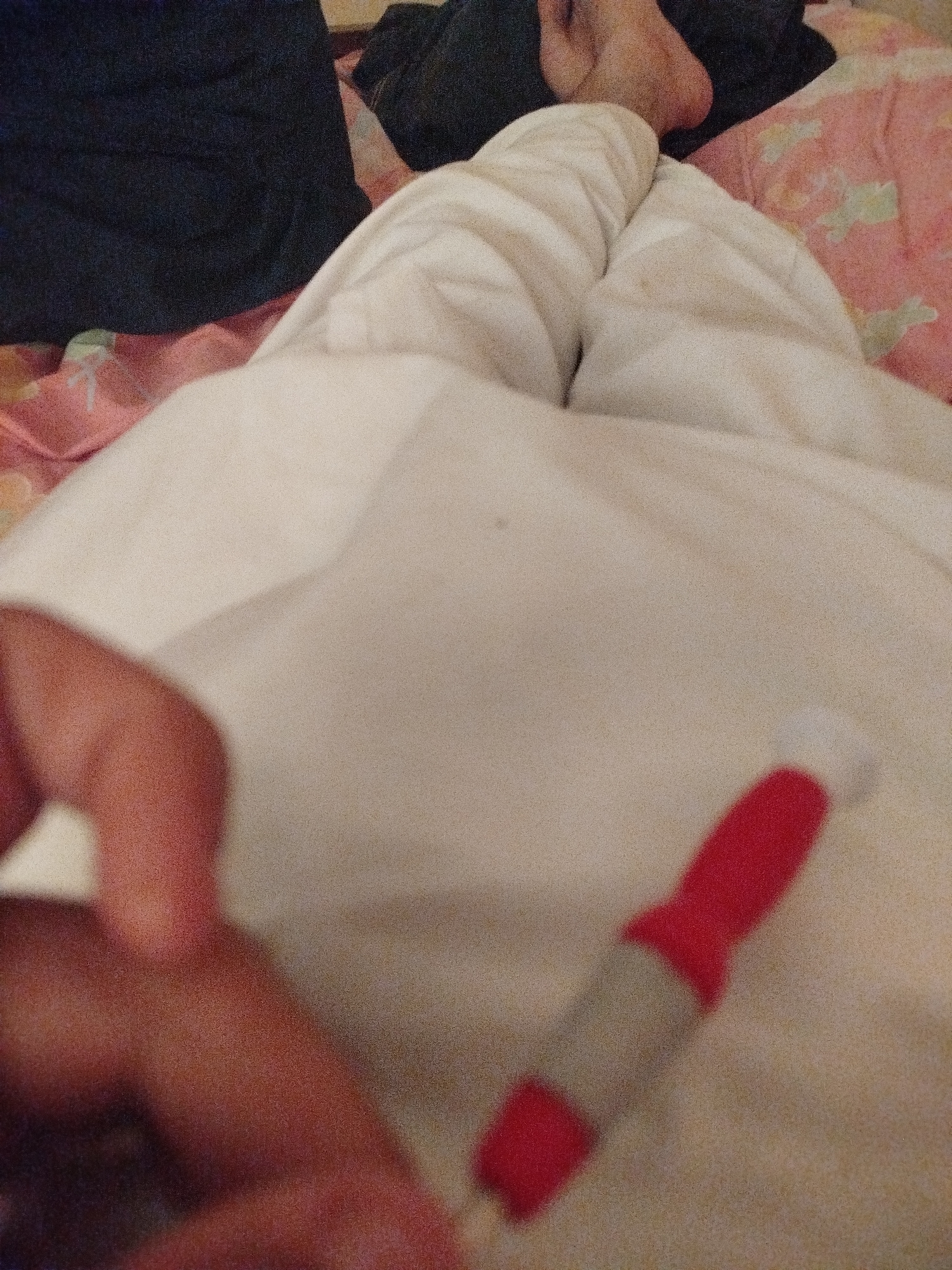 The image shows a close-up view of a person's legs covered with a white blanket. The person is wearing light-colored pants. Part of a hand is visible in the lower left corner, holding a red and grey object that appears to be a pen or a marker. The background shows a hint of a pink blanket with a floral pattern and dark fabric, suggesting a bedroom or a similar domestic setting. The lighting is dim, and the overall impression is of a relaxed or casual moment. There is no visible text in the image. The location context is Lassan, Pakistan. The activity appears to be resting or holding an object.