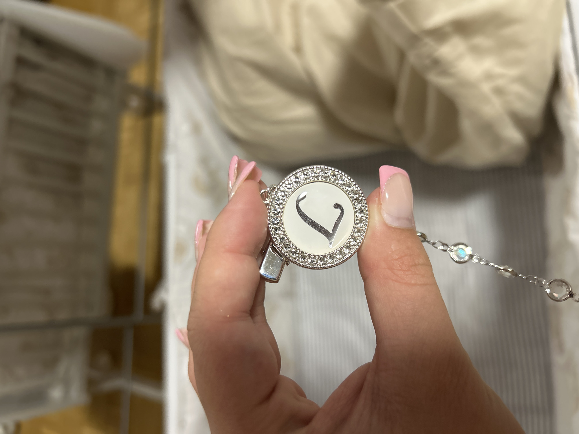 A person's hand with pink-painted nails is holding a round, silver-colored pendant. The pendant has a white face with a black letter "L" in the center. The edge of the pendant is decorated with small, clear stones. A delicate silver chain with additional small clear stones is attached to the bottom of the pendant. The background is blurred, but appears to be an indoor setting with furniture. This scene is not located in Waldbröl, Germany.