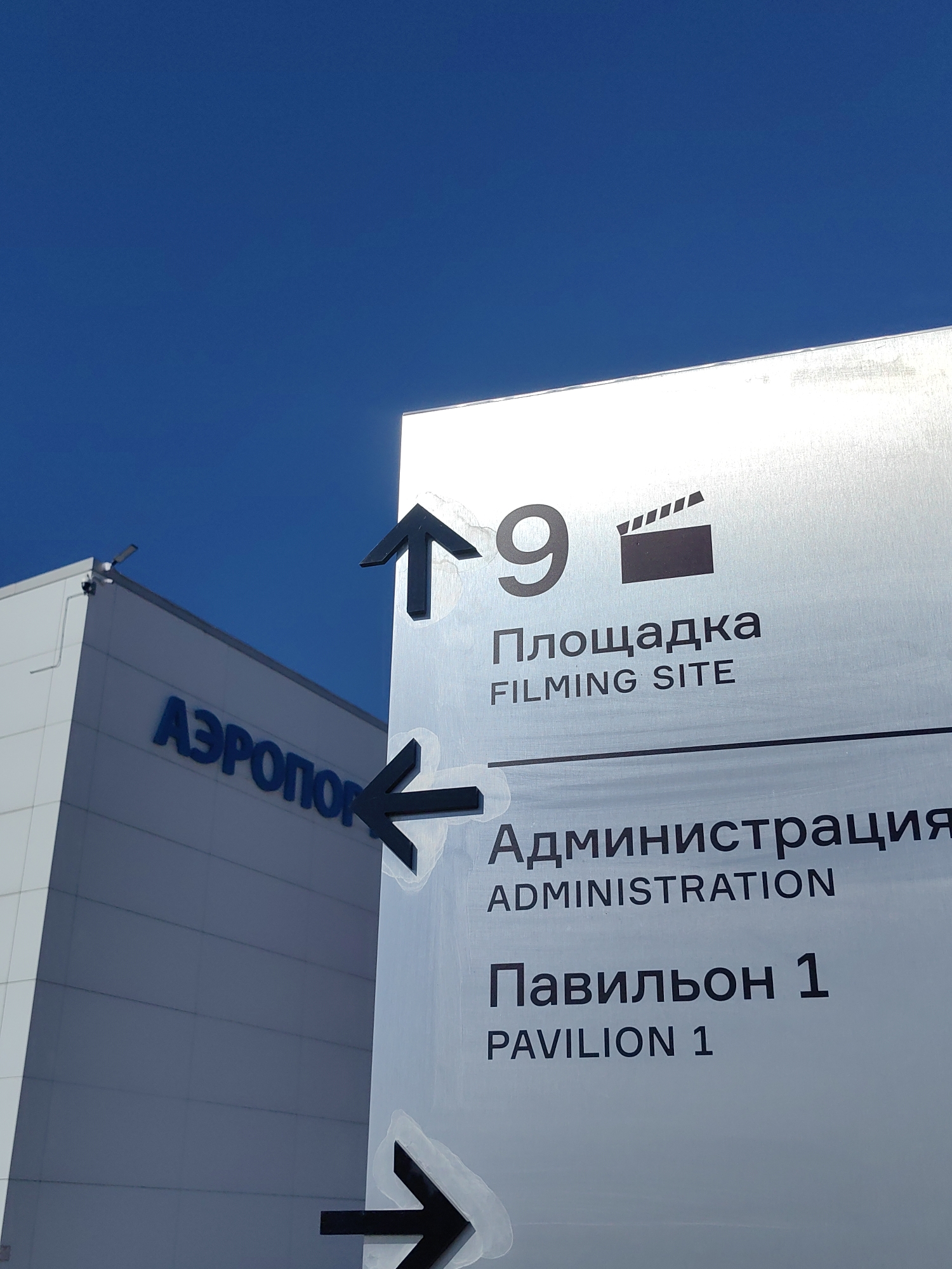This image features a close-up of a directional signpost with directional arrows and text in Russian and English. The sign is a light gray metallic material and has black text and arrows. The top section of the sign indicates "9" with a film clapperboard icon, followed by "Площадка" and "FILMING SITE". Below this is another section with "Администрация" and "ADMINISTRATION", and the final section reads "Павильон 1" and "PAVILION 1". Two black arrows are visible; one points upwards and slightly to the right, while the other points to the left.
In the background, a portion of a modern building with a light-colored facade and blue lettering that reads "АЗРОПОРТ" (AIRPORT) is visible, suggesting the location is an airport or related facility. The sky is a clear, vibrant blue. The image is taken from a low angle, looking up at the sign and building. The overall scene is clean and modern, with good lighting from the sun.