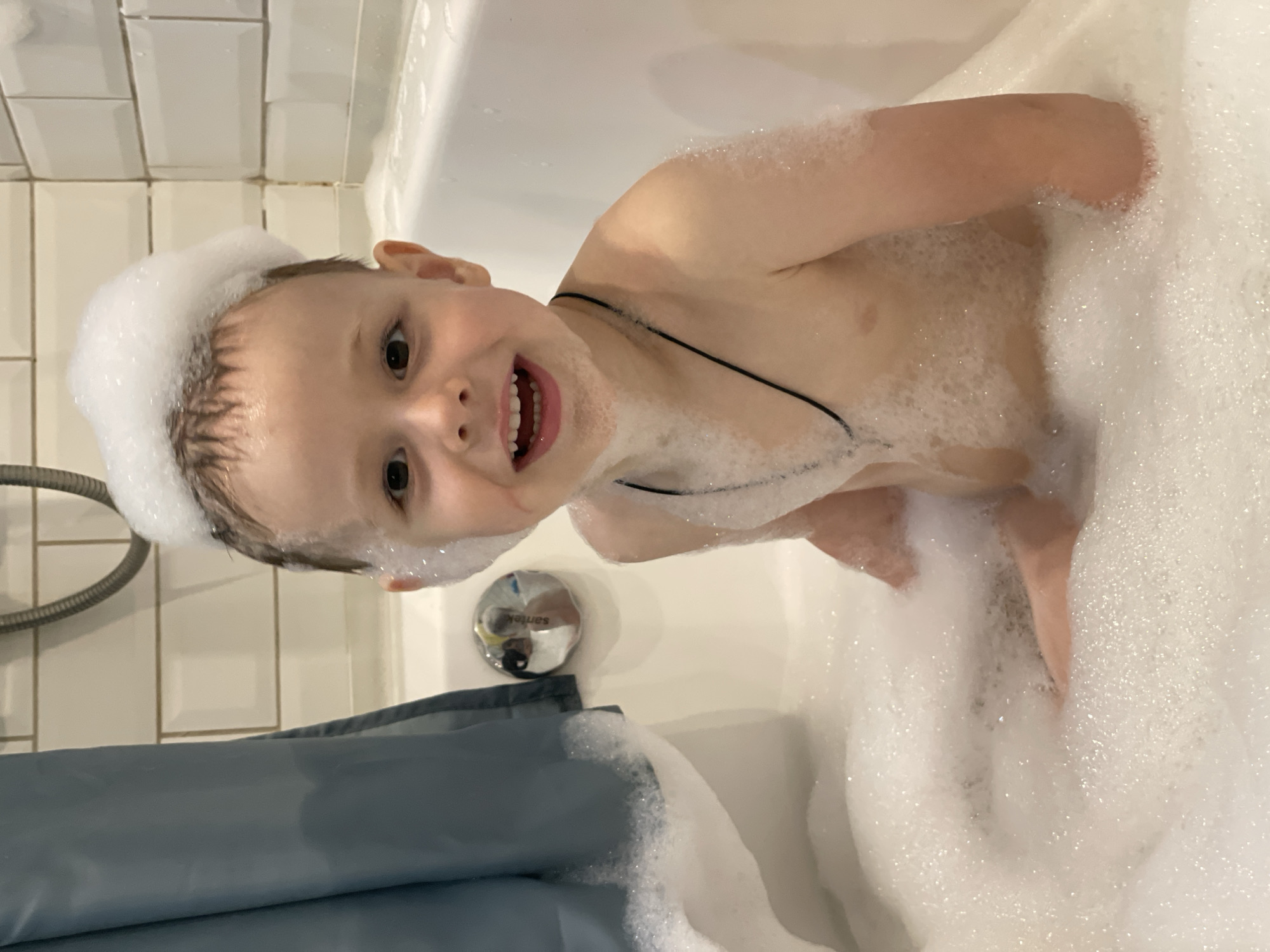 This media file shows a young child, likely a boy, in a bathtub filled with bubbles. The child is turned towards the camera with a wide, happy smile, revealing his teeth. He has bubbles on his head, creating a foamy hat, and his body is partially covered in suds. His skin appears fair and his hair is dark. He is wearing a thin black string or necklace around his neck.
The background shows a white tiled wall, typical of a bathroom. A gray shower curtain is visible in the lower part of the frame, and a metal shower head and hose are partially visible on the left. A chrome drain cover with the brand name "Sanitek" is also visible in the bathtub.
The scene is brightly lit, suggesting daytime, and the overall mood is playful and joyful, conveyed by the child's expression and the abundance of bubbles. The setting is clearly a bathroom, likely in a home, given the context of a child's bath. The location context of Izhevsk, Russia is provided externally and cannot be inferred solely from the image.