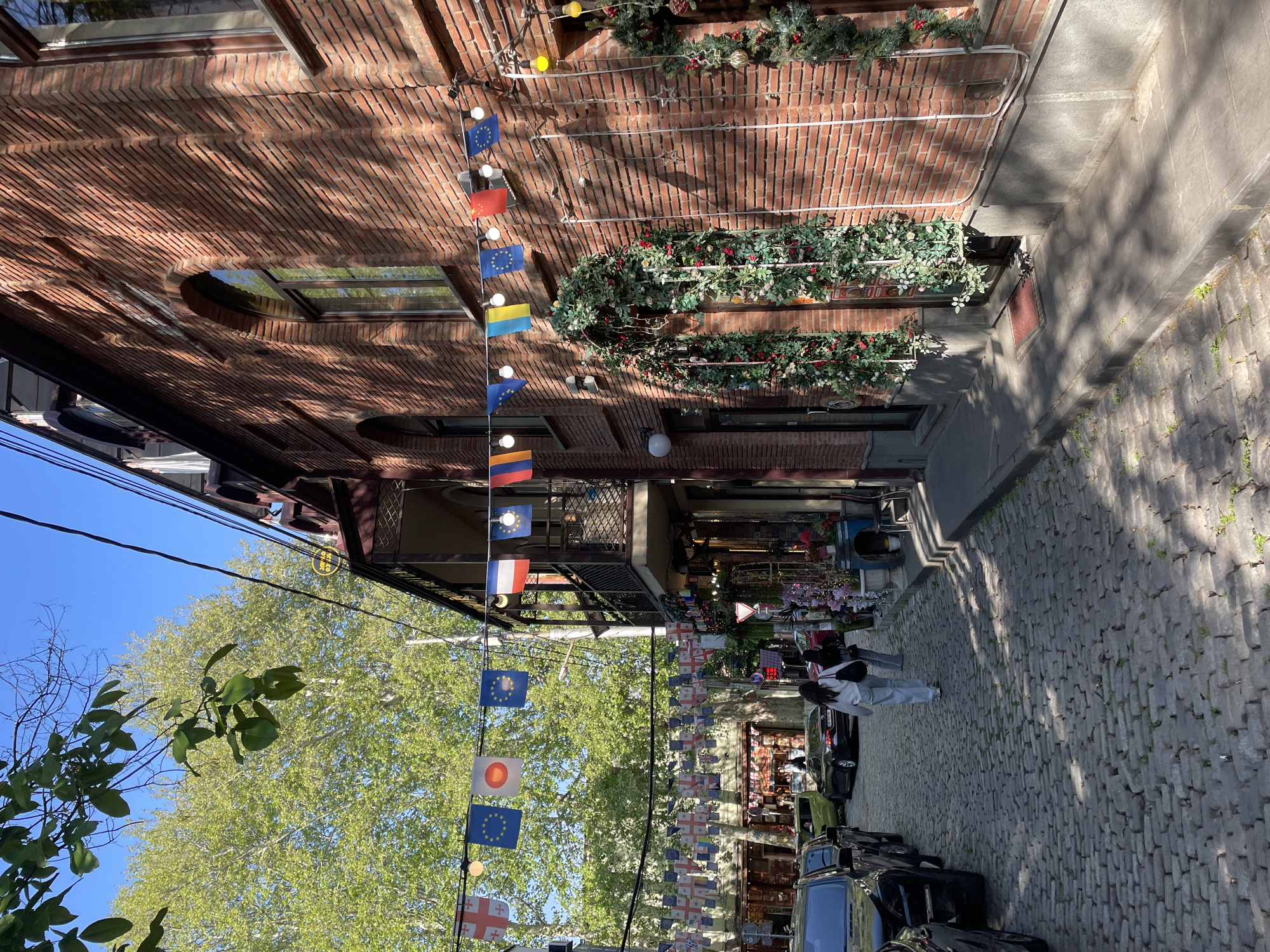 This media file captures a sunlit, charming street or alleyway in Tbilisi, Georgia, characterized by its cobblestone pavement and historic brick architecture. The image is taken from a slightly elevated perspective, looking down the street.
On the right side, a multi-story building of warm red brick features a mix of rectangular and a distinctive round window, reflecting the clear blue sky and surrounding greenery. The facade is festively adorned with garlands of evergreen foliage and red berries, along with string lights, suggesting a decorative or holiday theme. External conduits or wiring are visible along the brickwork.
Numerous small flags are strung across the street, creating a vibrant atmosphere. Identifiable flags include those of the European Union, Ukraine, Armenia, Poland, France, Japan, Georgia (with its red cross on white background), and England (St. George's Cross). A solid red flag, possibly representing China, is also visible among them.
Two individuals, appearing to be women, are seen walking on the cobblestone street towards the background. Several cars are parked along the left side of the street, and various shopfronts are visible at the base of the buildings.
To the left, large, green-leafed trees frame the clear blue sky. Bright sunlight casts distinct shadows on the cobblestones, indicating a clear, sunny day. A yellow circular sign with dark, illegible script, likely Georgian, hangs from a building on the far left. The scene evokes a lively and decorated urban space.