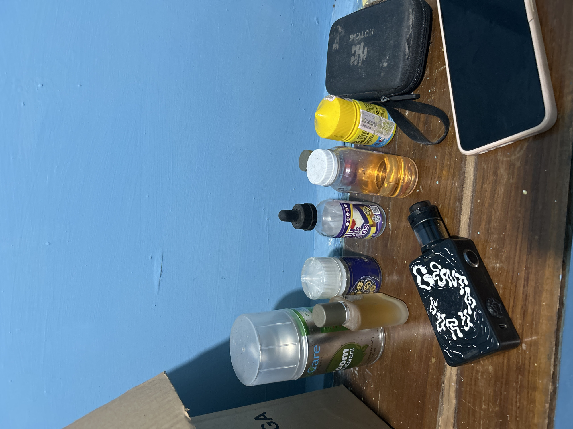 The image depicts a still life arrangement of various items on a wooden surface against a bright blue wall. In the foreground, a black electronic cigarette device with white swirling patterns is prominently displayed. To the right, a smartphone with a light-colored casing is partially visible, its screen dark. Behind these items, a collection of bottles and containers are arranged. These include a tall silver can labeled "Care" and "Boom Deodorant," a small clear glass bottle with a gold-colored liquid, a white-capped bottle with blue liquid, a clear bottle with an orange liquid and a black dropper top, a yellow-capped bottle, and a black zippered case. The background is a solid blue wall, which appears to be painted. The lighting suggests an indoor setting, possibly a room. There are no people or discernible actions taking place in the image. The scene does not contain any specific visual cues that would indicate a location within Purbalingga, Indonesia.