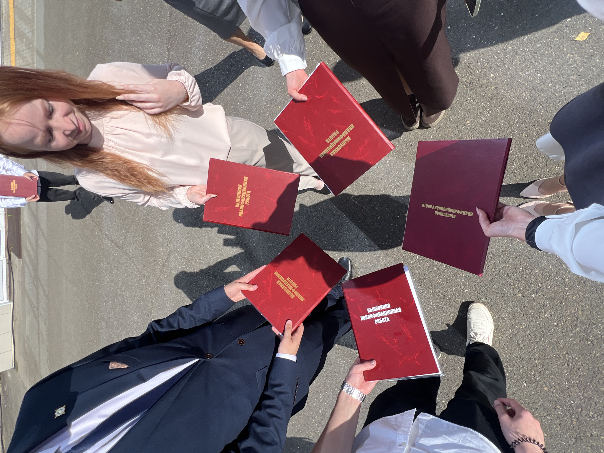 In an outdoor, paved area, likely in the city of Chaikovsky, Russia, a group of at least five individuals is gathered in a circular formation, each holding a red diploma or document. The documents have gold lettering on the cover, which reads "ВЫПУСКНАЯ КВАЛИФИКАЦИОННАЯ РАБОТА" (Graduation Qualification Work). The individuals are dressed in various semi-formal attire, including a dark suit, a light pink blouse, and white shirts. The lighting suggests a sunny day, with shadows cast on the ground. The ground appears to be asphalt, and there is a hint of a building or wall in the background, out of focus. The perspective is from a slightly elevated viewpoint, looking down at the group and their diplomas.