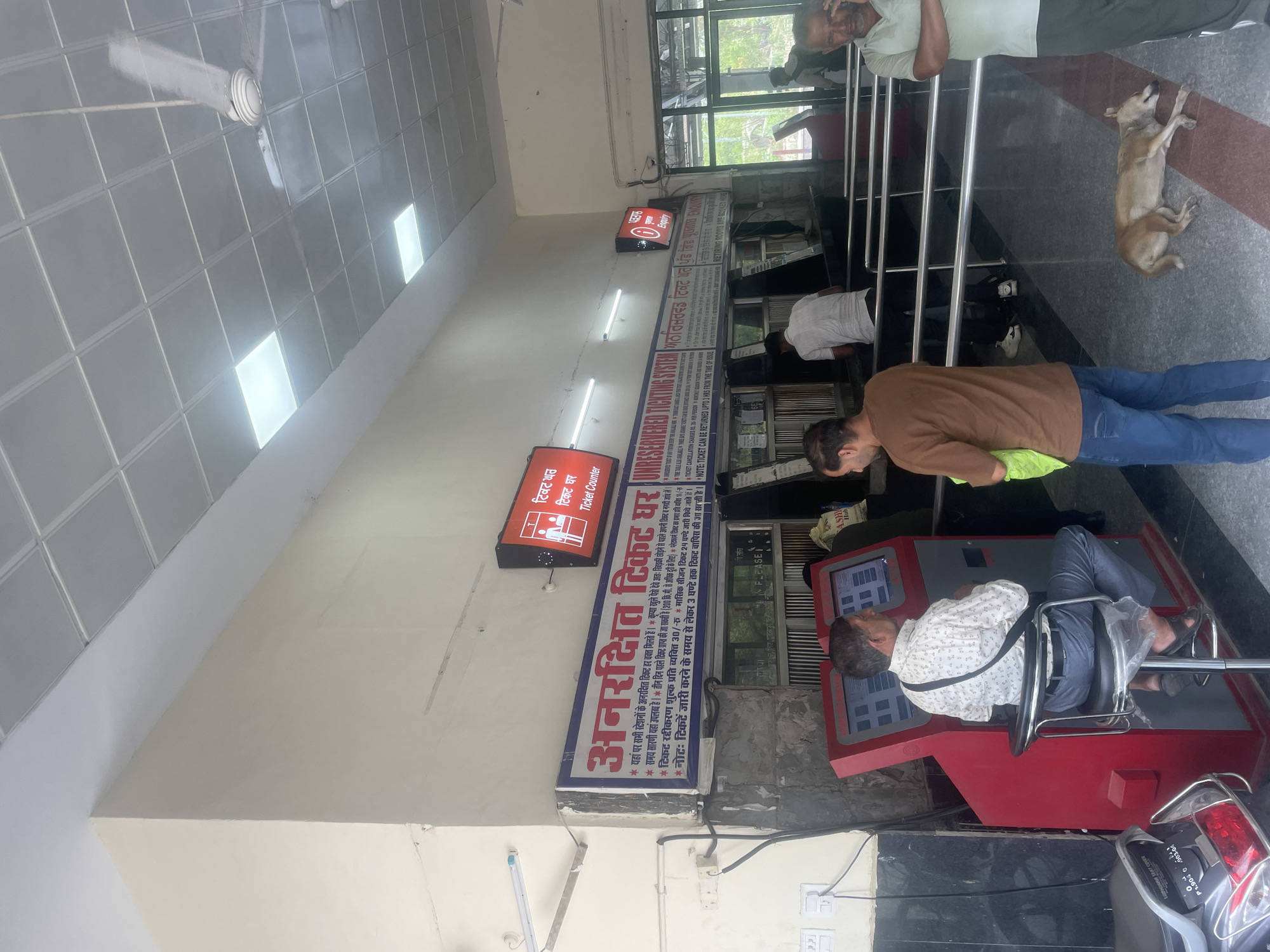 The image depicts the interior of a train station in Pathankot, India, specifically a ticket counter area. In the foreground, a man is seated on a stool in front of a red self-service ticket kiosk, appearing to interact with the screen. To the left, a motorcycle is partially visible. Behind the kiosk, a long counter with several windows is set up for ticket sales, with some individuals visible behind the counter and others in line or walking by. A sign above the counter indicates "TICKET COUNTER" in English and Hindi. Further in the background, there are more ticket windows and an "ENQUIRY" sign. A ceiling fan is visible, along with fluorescent lighting. The floor is tiled with a patterned design. A stray dog is lying on the floor in the middle ground, near a metal railing. The overall environment suggests a busy public transportation hub.