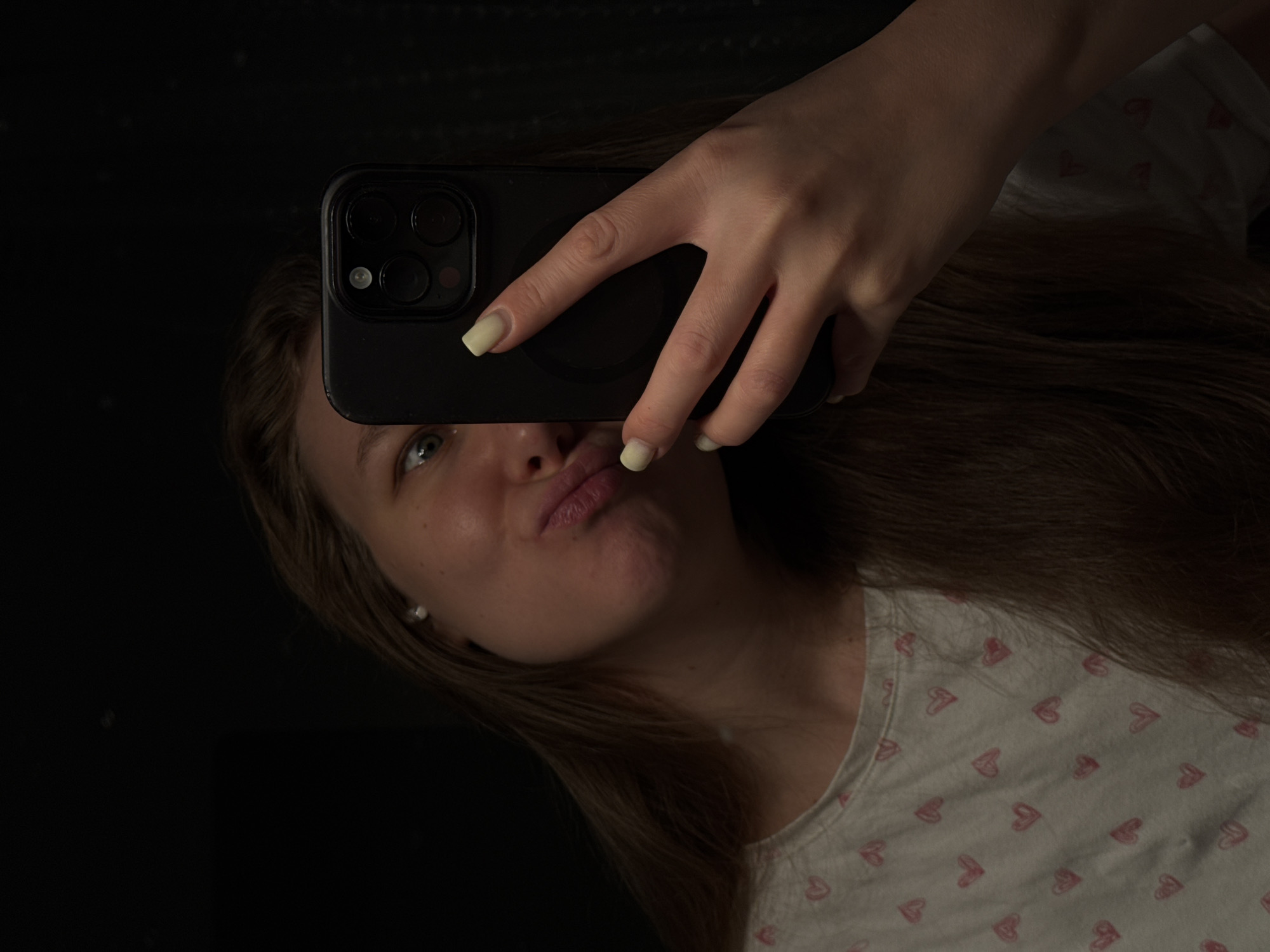 A selfie-style photograph shows a young woman holding a smartphone in her right hand. The smartphone partially obscures her face. She is wearing a white t-shirt with a heart pattern. The background appears to be a dark, possibly reflective surface, or a dark space. There is no visual evidence to indicate the location is Vladimir, Russia.