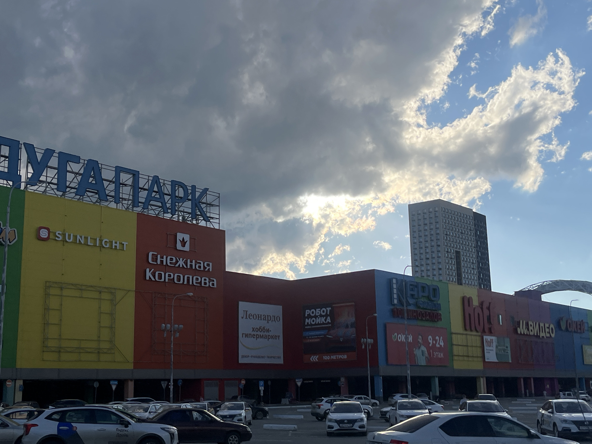 This is a wide shot of a large, multi-colored shopping mall named "YUG-PARK" in Yekaterinburg, Russia, under a dramatic sky with dark, heavy clouds on the left and bright sunshine with scattered clouds on the right. The mall's facade is divided into several large sections, each displaying different brand names. Prominently featured are "SUNLIGHT" in yellow and "СНЕЖНАЯ КОРОЛЕВА" (Snezhnaya Koroleva) in red. Other visible businesses include "Leonardo" (hobby hypermarket), "РОБОТ МОЙКА" (Robot Car Wash), "КЕРО" (KERO), "Hoff", and "М.ВИДЕО" (M.Video). A tall, dark residential building is visible behind the mall. In the foreground, a parking lot filled with numerous cars is seen, with some vehicles in motion and others parked. The overall impression is a bustling commercial area on a day with dynamic weather conditions.