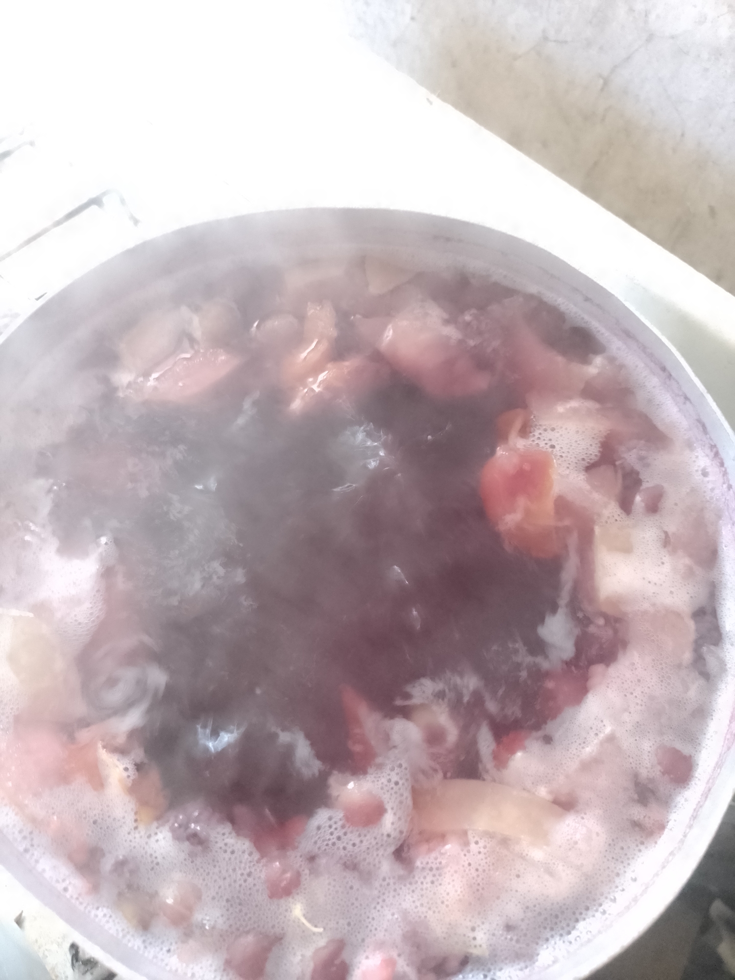 A close-up shot shows a white pot filled with a simmering reddish-purple liquid, likely a stew or soup. Small pieces of what appear to be food, possibly fruit or vegetables, are visible within the pot, some pinkish and others a darker shade. The surface of the liquid is covered in frothy bubbles and steam rises, indicating it is hot. The pot is sitting on a light-colored, possibly white, surface, and a portion of a white wall or countertop is visible in the background. The scene appears to be indoors, in a domestic setting.