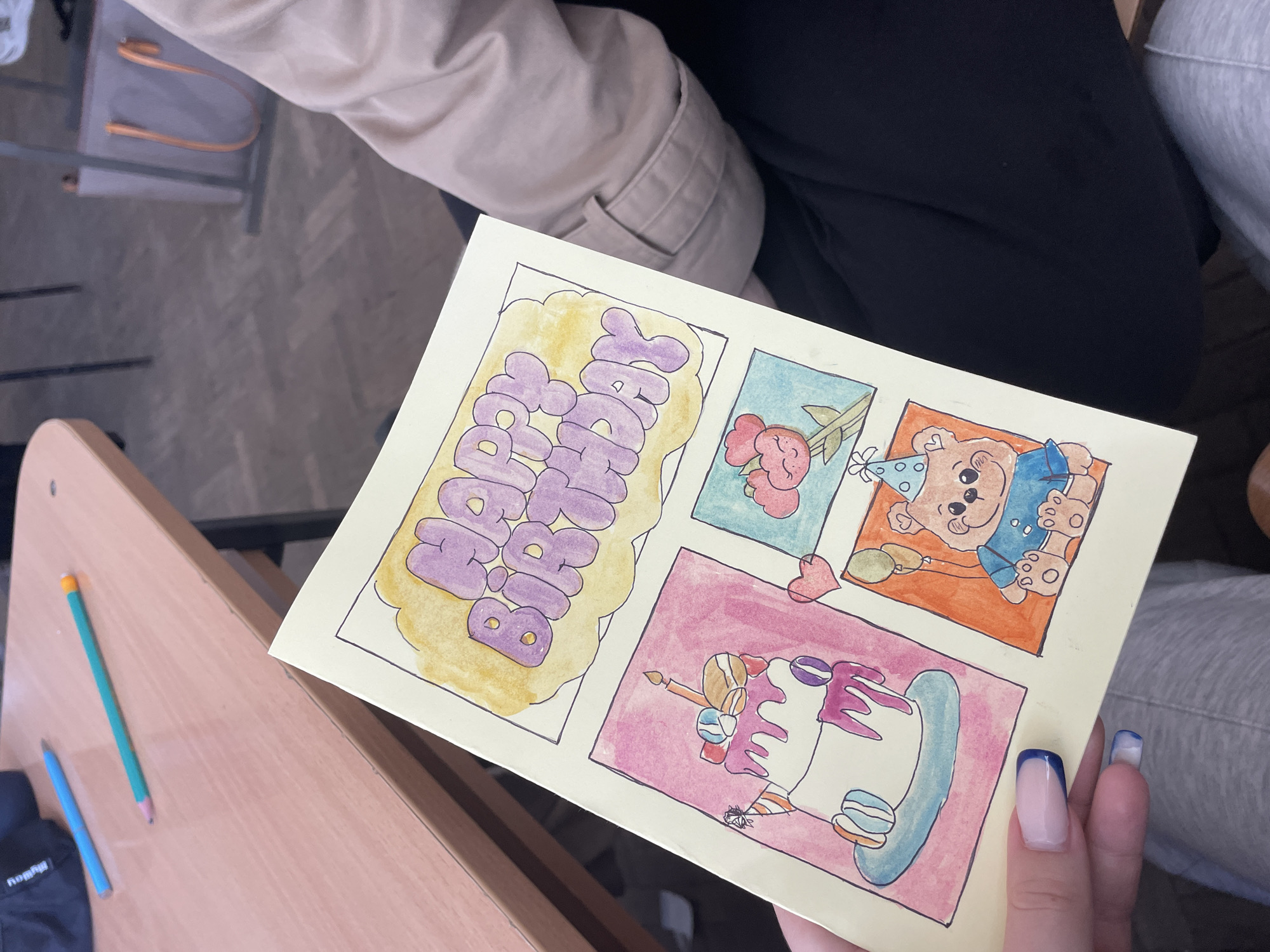 A birthday card is being held by a hand with manicured nails, the person wearing dark trousers and a beige jacket. The card has four sections: “Happy Birthday” written in stylized purple letters on a yellow background; a small drawing of two cartoon flowers and a heart on a blue background; a cartoon bear wearing a party hat and bib on an orange background; and a piece of cake with pink frosting and blue decoration, on a pink background. The card is being held above a wooden desk. Two pencils, one blue and one green, and a dark-colored wallet or case are visible on the desk to the left of the card. The setting appears to be inside, but the specific location within the city of Zlatograd, Bulgaria is not discernible.