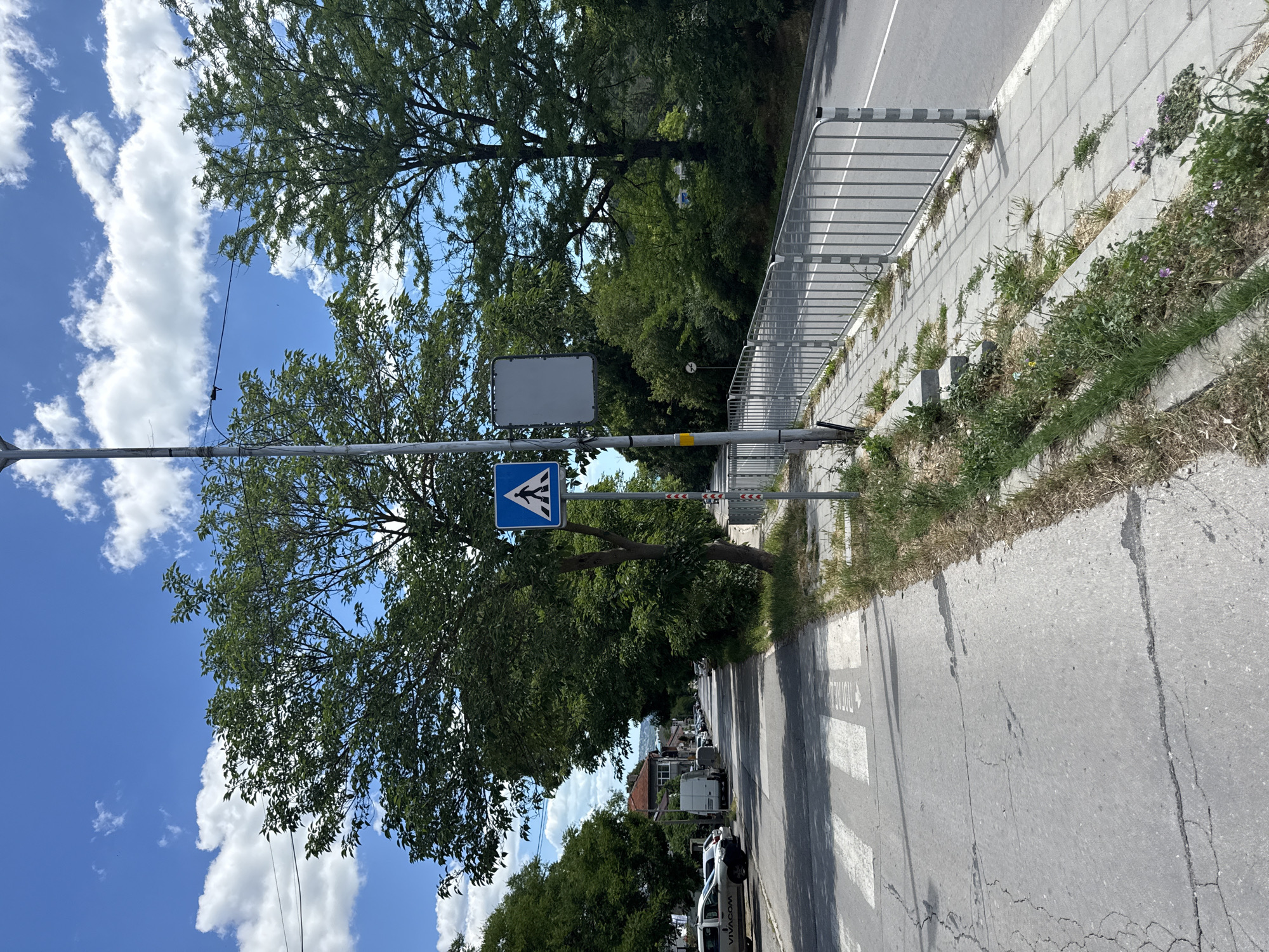 The image shows a street scene in an urban environment. A pedestrian crossing is visible on the left side of the road, marked with white stripes. A blue pedestrian crossing sign is mounted on a pole next to the crossing. A light pole with a blank rectangular sign attached stands near the pedestrian crossing sign. A metal fence runs along the right side of the road, separating it from a sidewalk. Trees line the street, providing shade. The sky is blue with scattered white clouds. Vehicles are parked along the street in the distance. The road surface appears to be asphalt, with some cracks visible on the pedestrian area. The environment appears to be a residential or commercial street with a mix of trees and buildings.