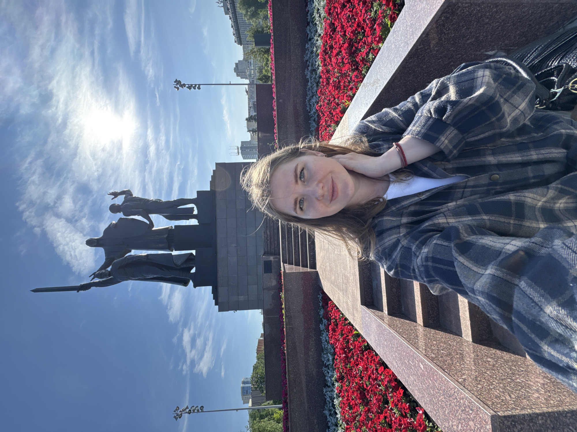 A young woman is featured in a selfie or portrait, taken outdoors in Perm, Russia, under bright daylight. She is positioned on the right side of the frame, looking towards the camera with a neutral to slightly smiling expression. She has light brown hair and wears a plaid button-up shirt over a white top, with a red bracelet on her left wrist.
Behind her, occupying the left side of the image, stands a substantial monument made of dark, polished stone. The monument features multiple bronze figures in heroic poses, with one figure holding a sword aloft.
In the middle ground, neat rows of vibrant red flowers and some silver/grey foliage are planted in elevated planters along the base of a granite structure. Beyond the monument and planters, urban buildings and trees are visible in the distance under a clear blue sky dotted with wispy white clouds. The bright sun is prominently visible in the upper left, causing some glare. The scene depicts a well-maintained public space, likely a memorial or significant landmark. No discernible text is visible on the monument or other elements within the frame.