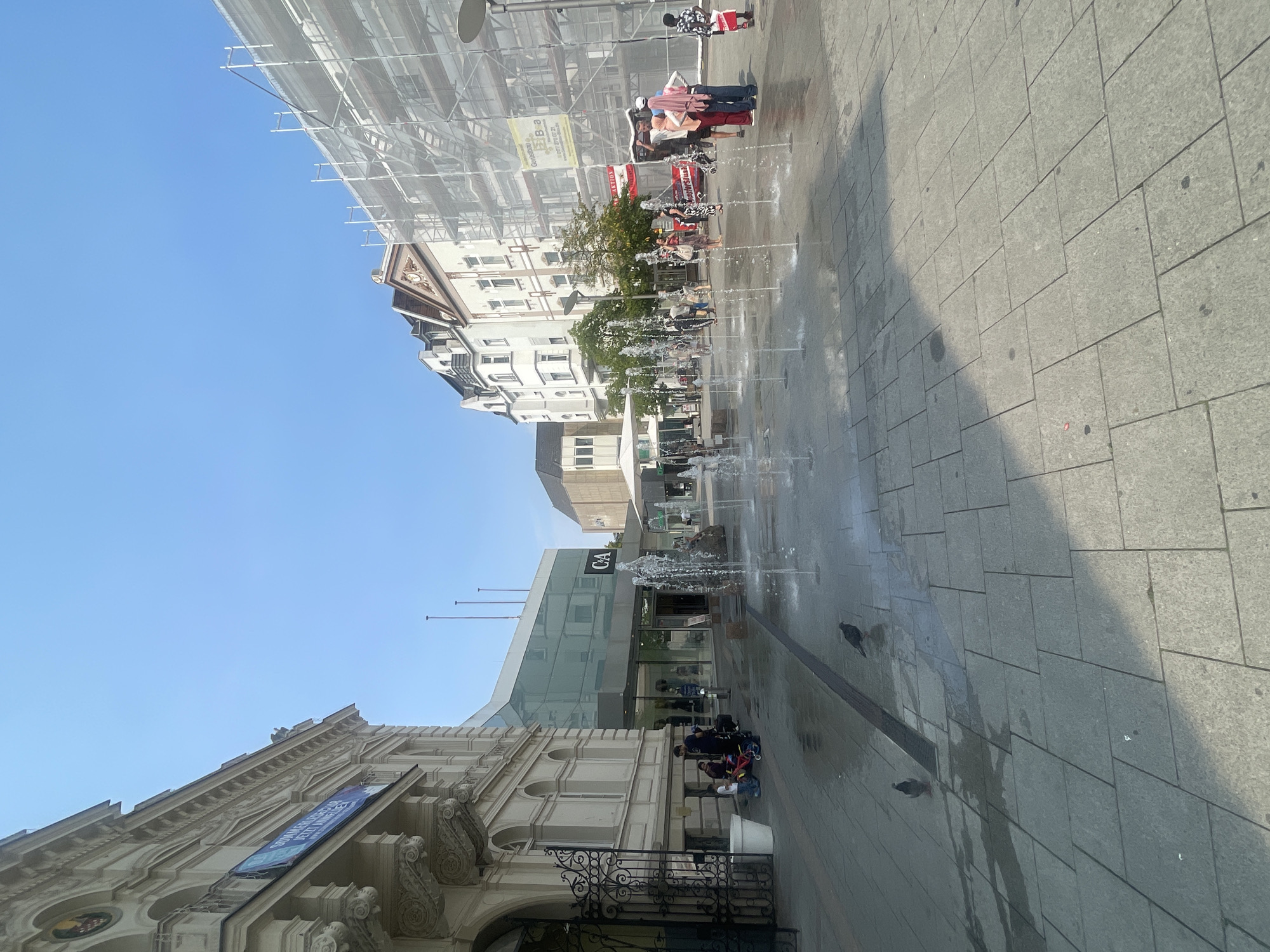 The image depicts a sunny daytime scene in the city of Iserlohn, Germany, likely a public square or pedestrian zone, featuring a C&A store. In the foreground, a modern fountain system with multiple jets of water is active, spraying water onto the paved ground. Two pigeons are visible on the wet pavement. To the left, an ornate, historic building with classical architectural elements stands beside a modern glass-fronted structure housing the C&A store. Several people are gathered in front of the C&A entrance, and a family with young children is near a child's tricycle. Further into the scene, more people are walking and conversing. On the right side of the image, a building is covered in scaffolding and white protective netting, indicating ongoing construction or renovation. The sky is clear and blue, suggesting good weather. The overall atmosphere is that of a bustling urban commercial area.