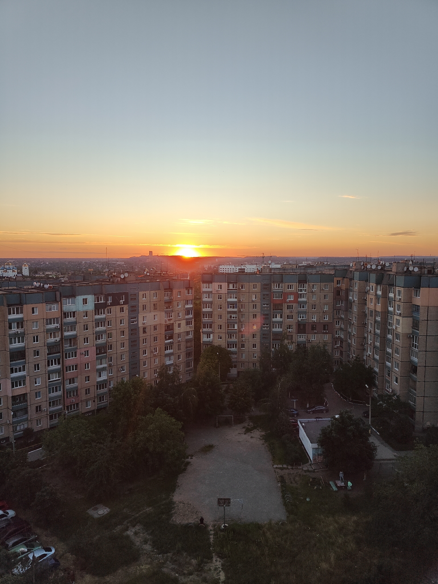 This is a high-angle, medium shot of a residential area in Kryvyi Rih, Ukraine, at sunset. The sun is low on the horizon, casting a warm orange glow across the sky and illuminating the tops of several large, multi-story apartment buildings. The buildings are constructed from concrete and brick, with numerous windows and balconies. In the foreground, a patch of overgrown grass and trees separates the buildings from a small, paved courtyard area. A basketball hoop and backboard are visible in the courtyard, along with a small group of people, including a child, gathered around some playground equipment. Several parked cars are visible on a patch of grass to the left of the frame. The overall impression is one of an ordinary urban neighborhood during the evening hours.