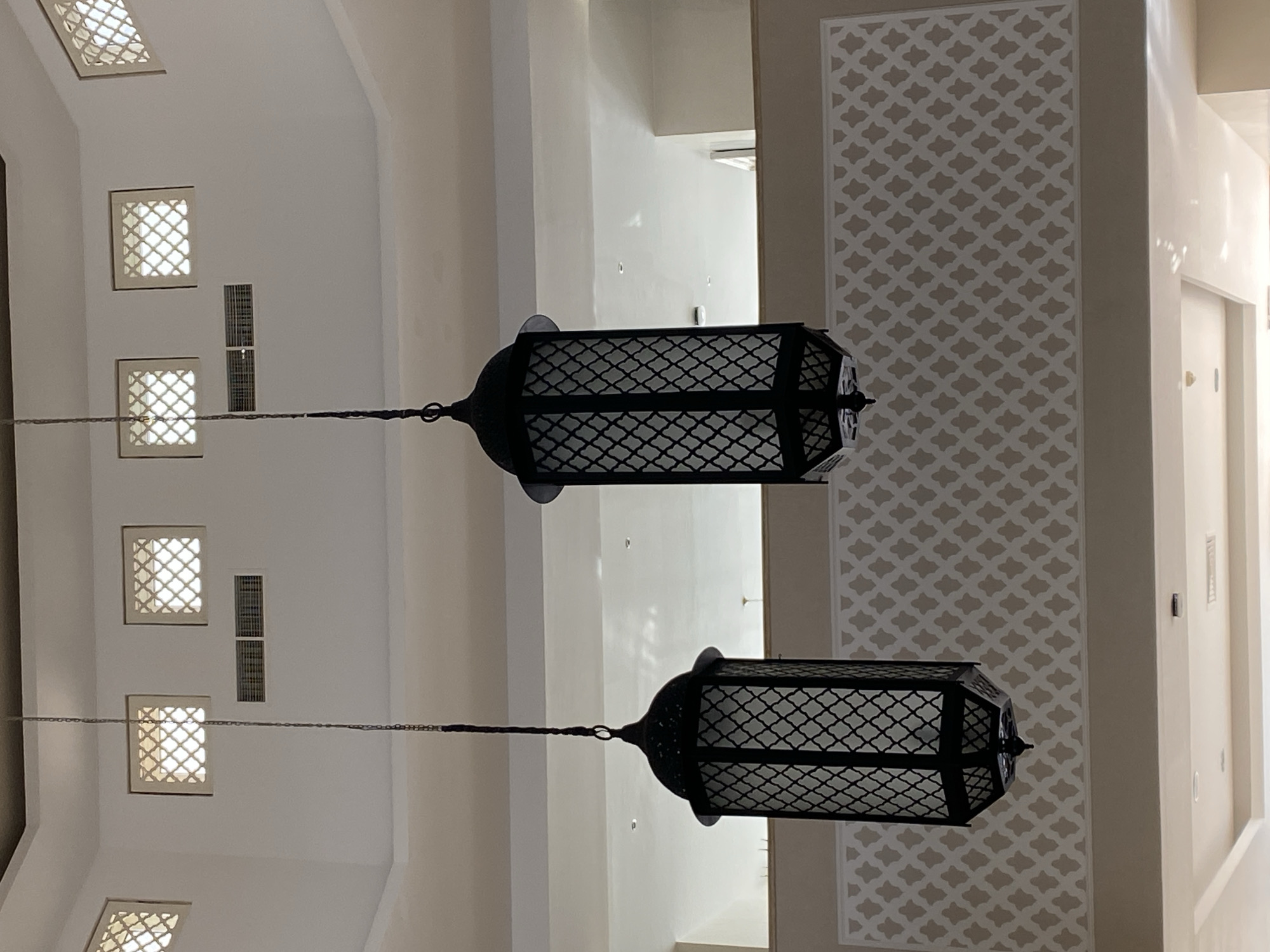 The image shows two ornate, black metal lanterns suspended from the ceiling by chains. The lanterns have a lattice-like design with white glass panels. Above the lanterns, the ceiling is white and features square windows with decorative grilles. A patterned frieze runs along the upper portion of the walls. The overall impression is one of interior architecture, possibly in a hotel or public building. There are no people or actions depicted in the image. The scene is likely located in Sharm El Sheikh, Egypt, given the architectural style and the mention of the city.