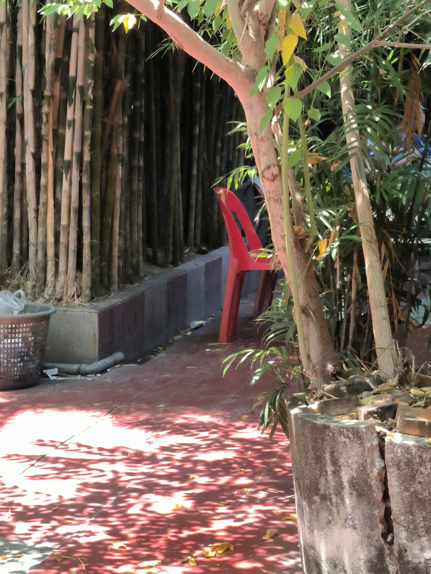 The photograph, taken in Lhokseumawe, Indonesia, shows a red plastic chair positioned near a bamboo fence. The chair sits on a red tiled patio area, dappled with shadows from overhead foliage. A tree with slender trunks and green leaves grows nearby. A grey plastic trash basket is visible to the left of the frame. A low concrete border with a dark grey top runs along the bamboo fence. Litter is scattered on the patio surface. A dilapidated concrete planter, partially broken, is situated on the right side of the frame. The bamboo fence consists of numerous closely packed, light brown bamboo stalks.
