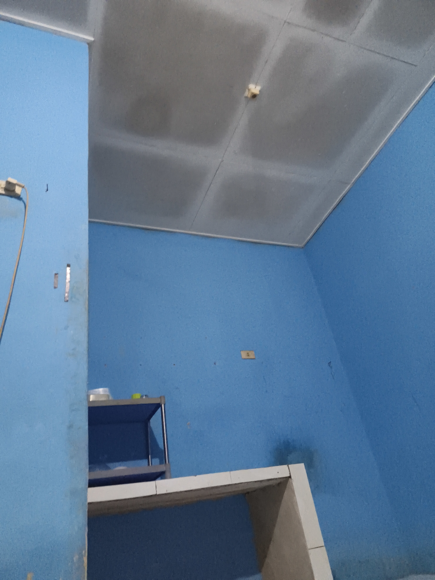 The image shows an interior view of a room with blue walls and a gray drop ceiling. On the left, a portion of a blue wall is visible, with a beige electrical cord hanging down and connected to a wall outlet. Below the outlet, there are two rectangular openings in the wall. In the center of the image, a white shelf unit with blue shelving is placed against the blue wall. The shelf unit appears to be holding a few white and green objects. A white desk or table is positioned in the foreground, extending from the left side of the image. The room appears to be unfinished or under renovation, with visible seams and joints in the ceiling and walls. The lighting is dim, casting shadows in the room. The scene is consistent with a residential or storage space in Pangkalpinang, Indonesia, possibly an unfinished room.