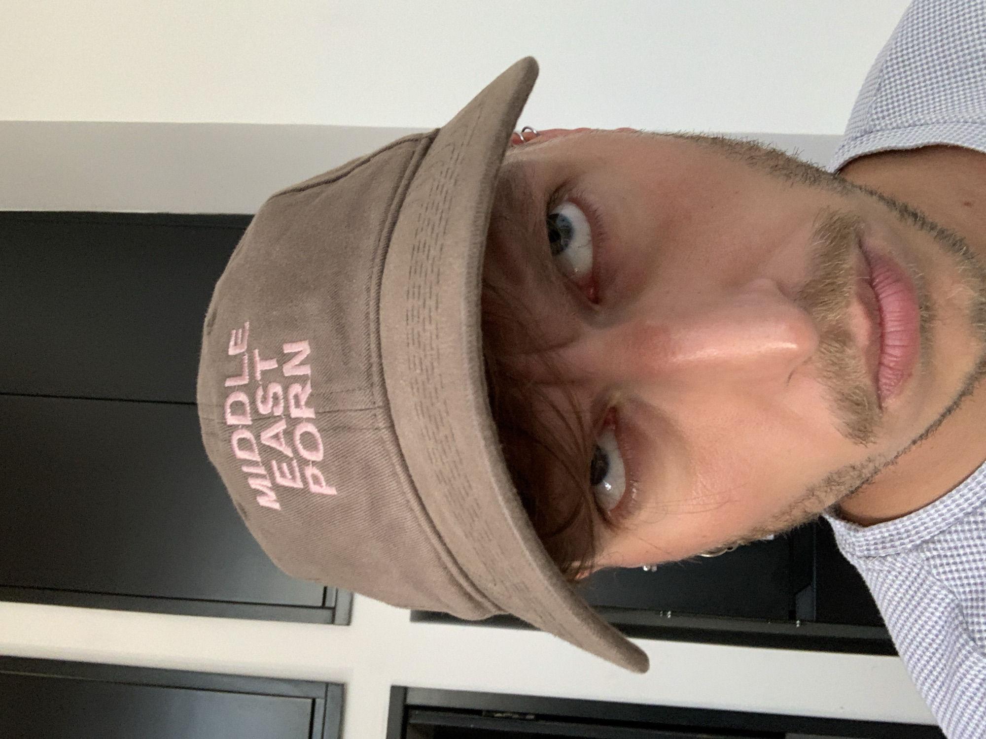 A man with a beard and light eyes is wearing a brown baseball cap with the words "MIDDLE EAST PORN" embroidered in pink. He is also wearing a light blue checkered shirt. The background consists of a white wall with a dark cabinet. The location, Tel-Aviv, Israel, is not discernible from the image.