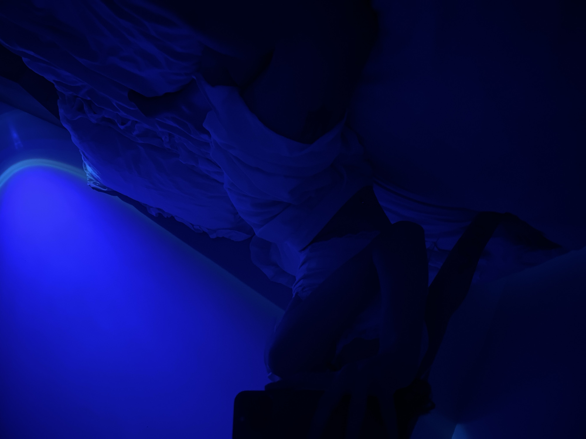 A dimly lit bedroom scene, bathed in a deep blue hue from an unseen light source. A person is lying on a bed covered with rumpled white linens. The person is seen in silhouette, lying on their side, with their head resting on a pillow and their hand holding a mobile phone. The person appears to be wearing a white garment. The environment suggests a private indoor space, possibly a bedroom, with a wall and ceiling visible in the background, also illuminated by the blue light. No specific details relating to the city of Ternopil, Ukraine, are discernible from the visual information provided in the image.