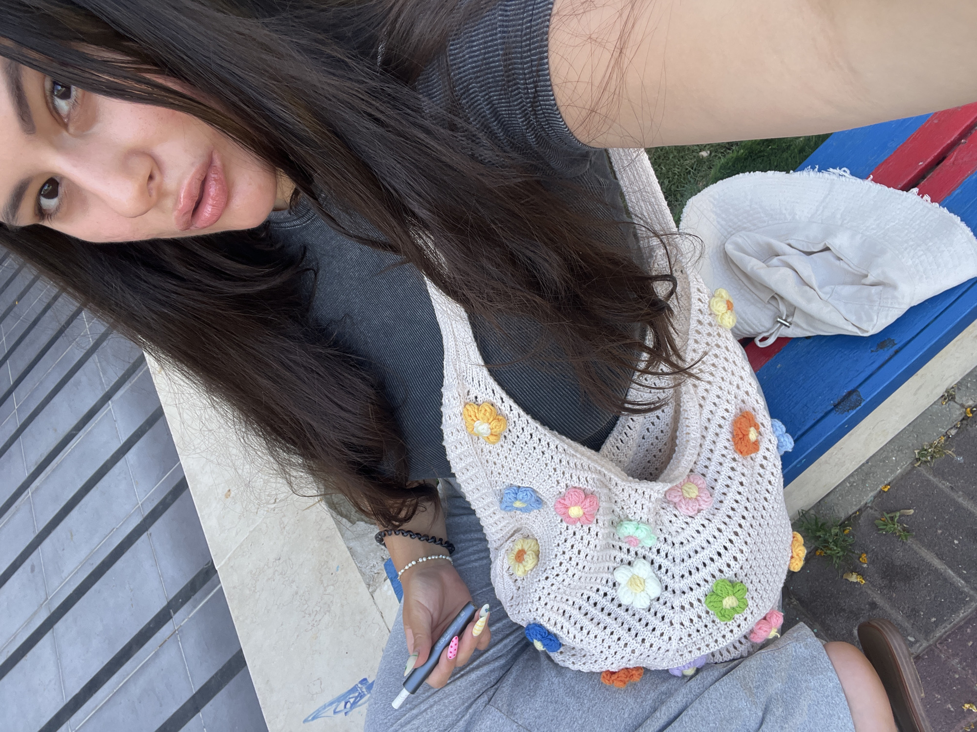 An overhead, medium shot captures a young woman sitting on a public bench in Kiryat Bialik, Israel. She is wearing a grey t-shirt and grey sweatpants, and her dark hair cascades around her shoulders and torso. In her left hand, she holds a vape pen. Draped across her lap is a light-colored, crocheted bag adorned with colorful floral appliques. To her right, resting on the bench, is a white sun hat. The bench itself is painted with red and blue stripes. The background reveals a tiled walkway and a patch of green grass, indicative of an outdoor urban setting. The lighting suggests it is daytime.