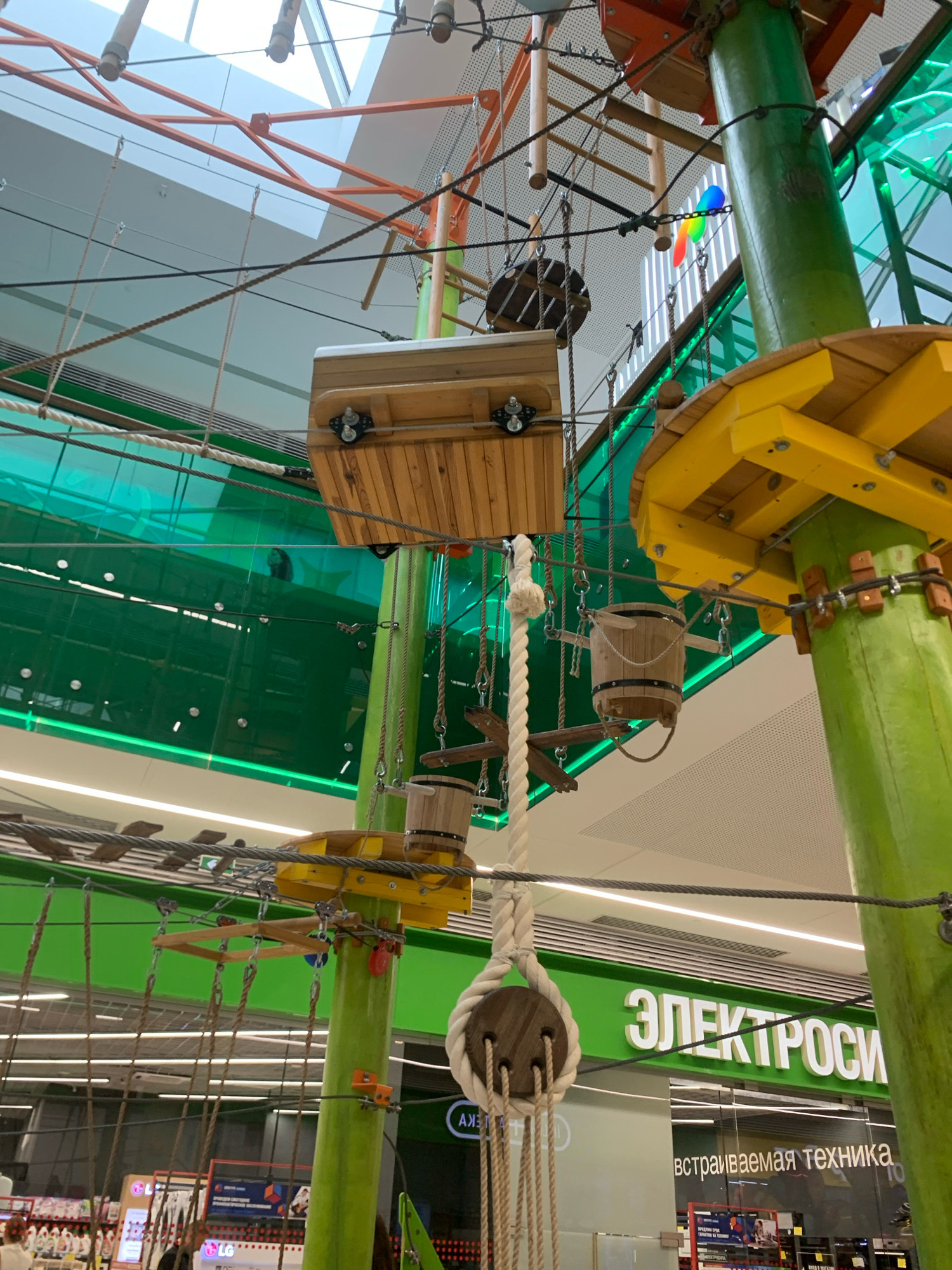The image shows an indoor rope park, possibly in a shopping mall, with various wooden platforms, ropes, and climbing elements suspended from a ceiling. In the background, a large green sign with white Cyrillic lettering reads "ЭЛЕКТРОСИ," which translates to "ELECTROSITY" or "ELECTROSILA." Below this sign, in smaller Cyrillic text, it says "встраиваемая техника," meaning "built-in appliances." This indicates the location is likely an electronics store in Minsk, Belarus, as "ELECTROSILA" is a known chain of electronics stores in Russia and Belarus. There are no people clearly visible in the foreground, but a shadowy figure can be seen through the green glass barrier in the middle ground. The overall environment appears to be a public indoor space with high ceilings and bright artificial lighting.