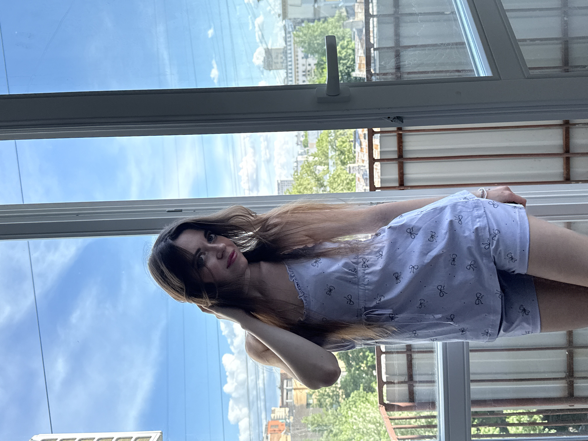 A woman is standing by an open window. She is fair-skinned with long, brown hair and is wearing a light-colored pajama set with a small print. The background is a view of a city skyline, including buildings and greenery. The city is Moscow, Russia. The window is open and the sky is visible. A metal balcony is visible outside the window.
