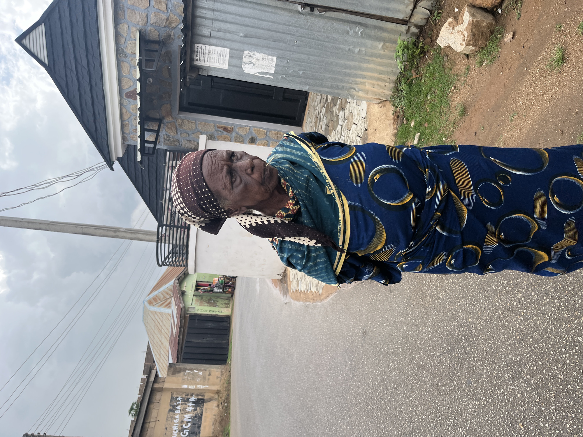 An elderly Black woman is standing on a paved road in Jos, Nigeria. She is wearing a blue patterned dress with yellow accents and a brown and white head covering. The woman is looking towards the left of the frame. Behind her is a building with a combination of corrugated metal, stone, and painted surfaces. In the background, there are other buildings and a cloudy sky. Power lines stretch across the top left portion of the frame. The road appears to be paved asphalt. Graffiti is visible on a wall on the left side of the road.