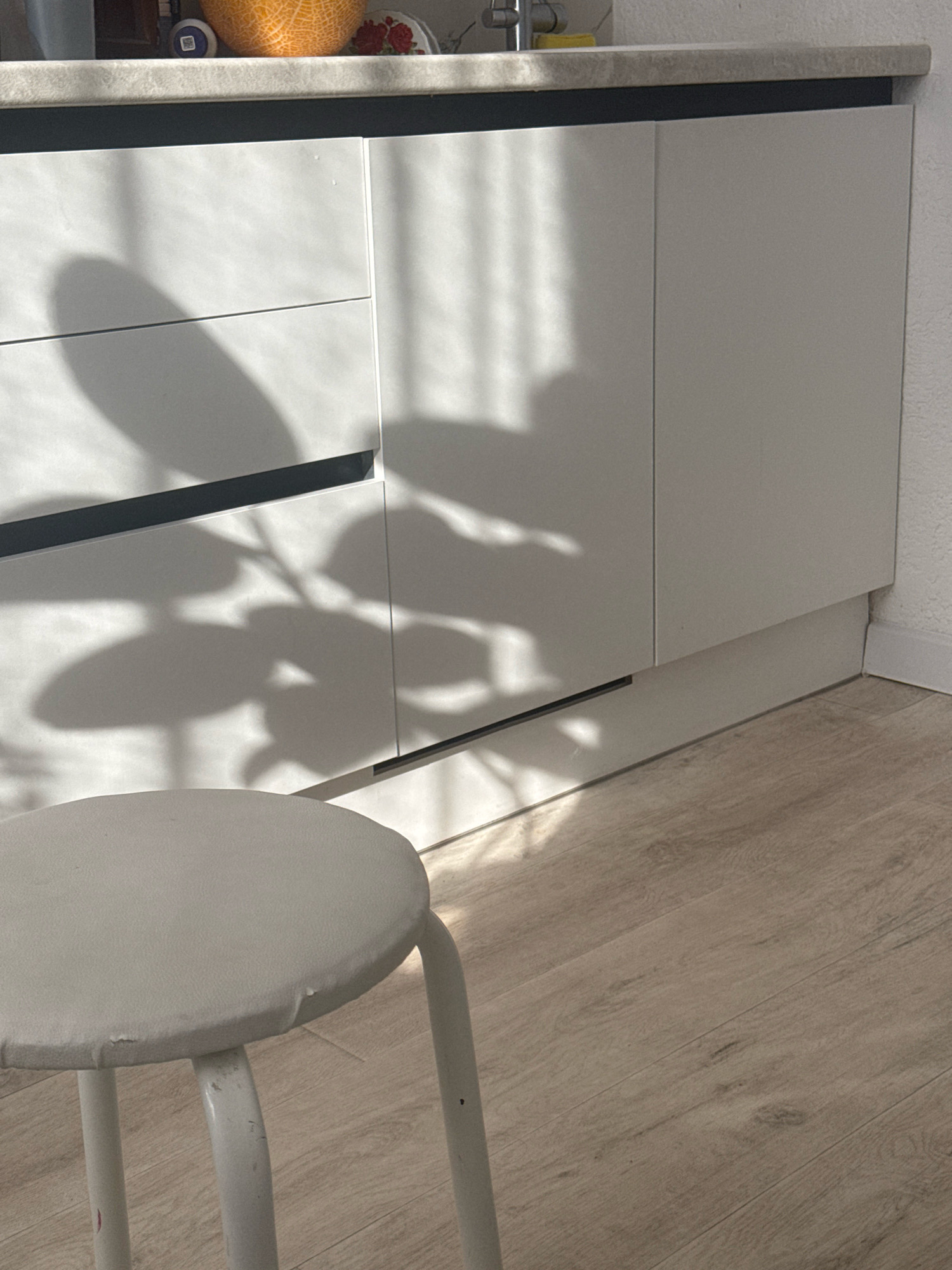 This is an eye-level, medium shot of a corner of a modern kitchen, possibly in Ufa, Russia. The foreground features a white stool with a circular seat and three thin legs. The stool is positioned on a light brown, wood-plank floor. In the background, there are white kitchen cabinets with thin, dark horizontal handles. A shadow of a plant with large leaves is cast across the cabinets and a portion of the floor. Sunlight streams in from the left, creating vertical light patterns on the cabinets and highlighting the wood grain of the floor. The countertop is a textured, light gray material. On the countertop, an orange, netted object, possibly a melon, is partially visible. The overall impression is one of a clean, contemporary kitchen with natural light enhancing the textures and shapes within the space. There are no people or animals visible in the image.