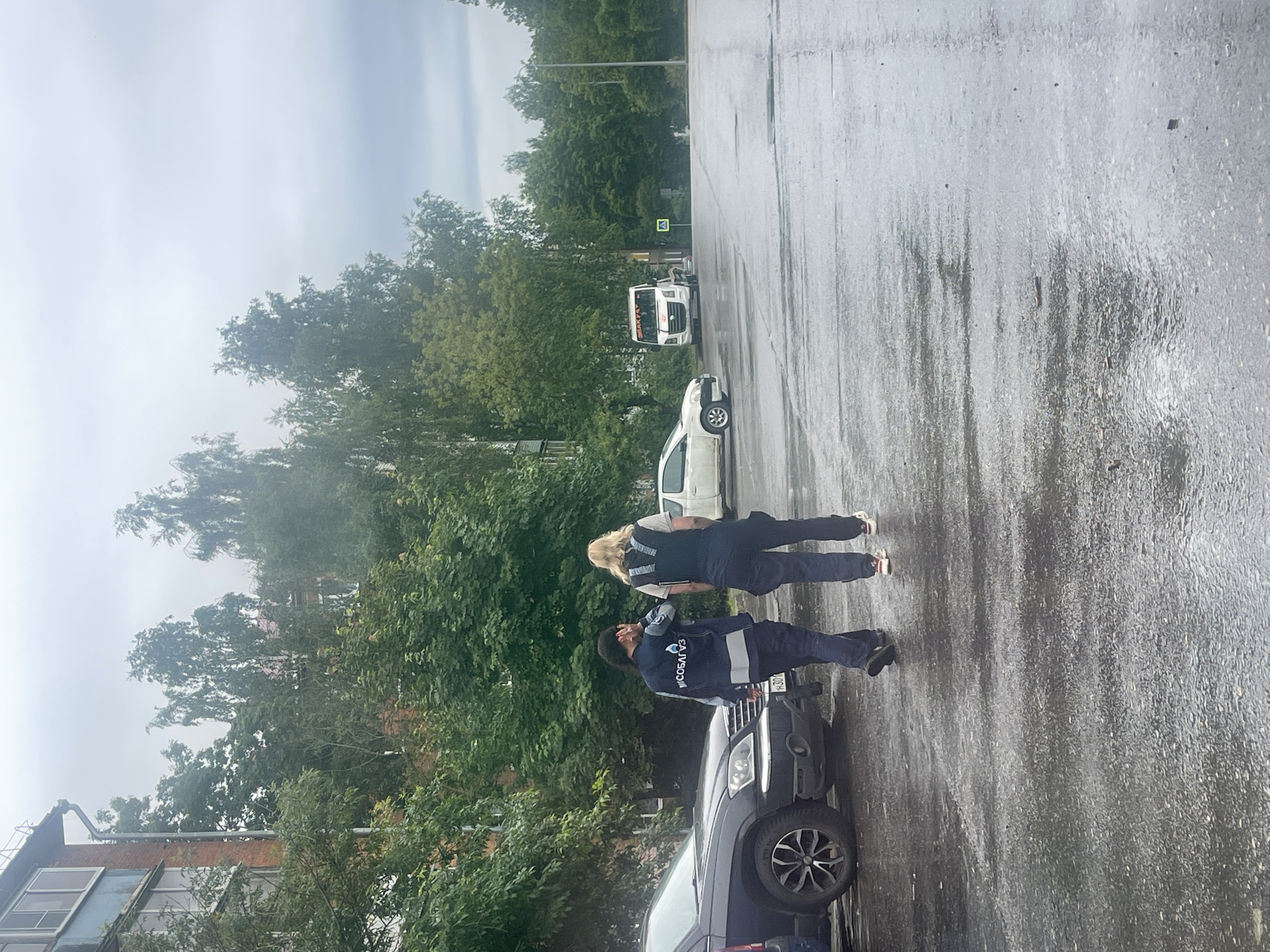 Two individuals, one wearing a dark blue jacket with reflective stripes and the text "МОСОБАЛГАЗ" on the back, and the other in a dark vest over a lighter shirt, are walking away from the camera on a wet asphalt street in Shakhovskaya, Russia. The person in the blue jacket has dark hair and is reaching up towards their head, while the person in the vest has blonde hair. A dark colored SUV is parked on the left side of the street. In the background, there are trees, a multi-story brick building, a white truck, and a yield sign. The sky is overcast.