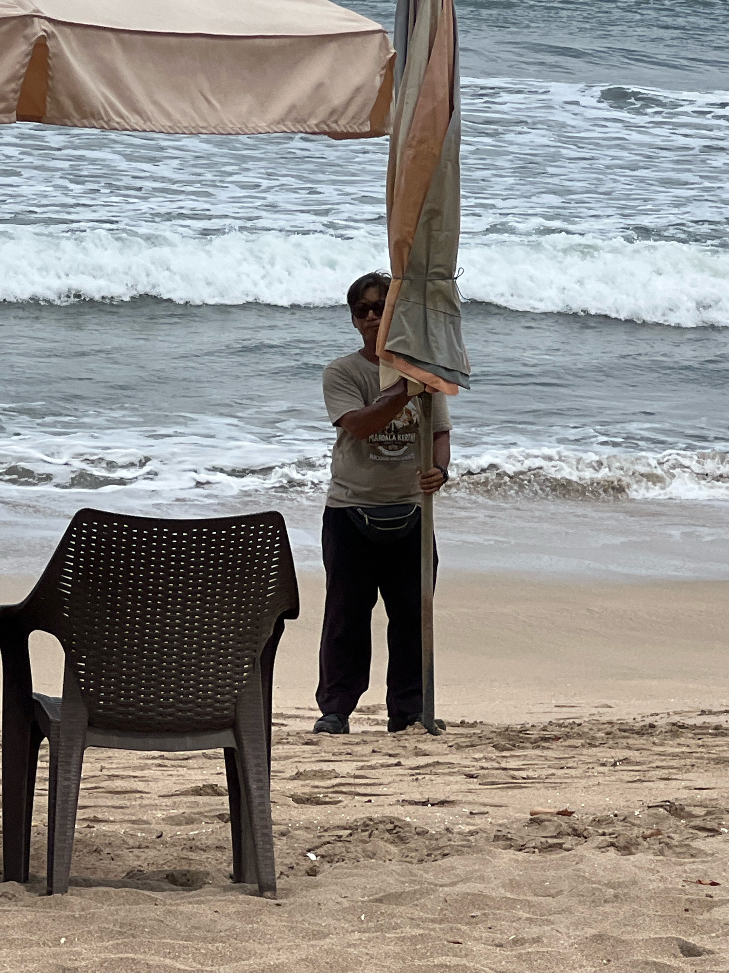A man stands on a sandy beach in Kuta, Indonesia, holding a folded-up beach umbrella. He is wearing a light-colored t-shirt with text and graphics, dark pants, sunglasses, and a dark fanny pack around his waist. The umbrella is a light tan and grey color, and it is tied closed with a rope. In the foreground, a dark brown plastic beach chair faces away from the camera. The ocean waves are breaking in the background, and a large beach umbrella is partially visible overhead. The sand is damp and shows tracks and small debris.