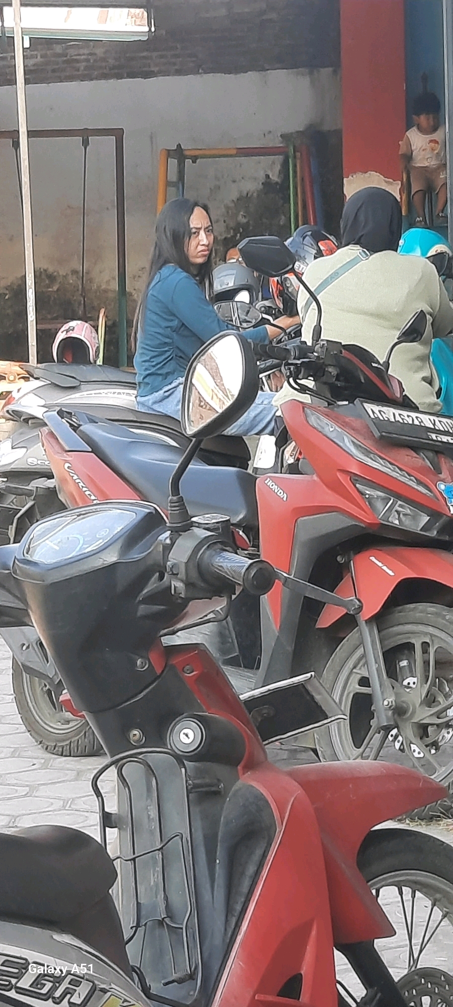 In Selopuro, Indonesia, a woman with long dark hair, wearing a teal long-sleeved shirt and blue jeans, sits on a red motorcycle. She appears to be looking to her left with a concerned expression. To her right, another person wearing a light-colored top and a hijab is seated on a motorcycle. In the background, a young child sits on a colorful structure, possibly part of a playground. The scene is set outdoors, with motorcycles parked in what appears to be a street or parking area. The environment includes brick walls and a tiled pavement.