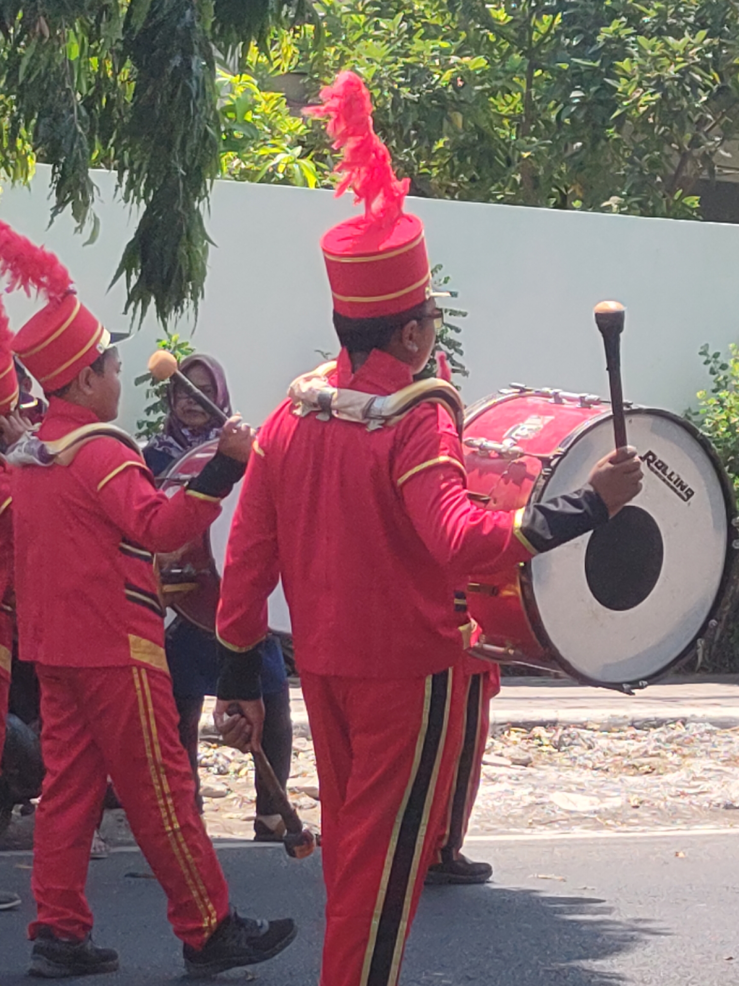 In Ngasem, Indonesia, a marching band procession is underway. Two young boys, dressed in red uniforms adorned with gold trim and bright red feather headdresses, are prominent. The boy on the left is marching forward, while the boy on the right carries a large bass drum. He is holding a drumstick and appears to be in mid-action, ready to strike the drum. Behind them, partially visible, a woman in a headscarf observes the proceedings. The background consists of lush green foliage and a white wall, suggesting an outdoor urban or suburban setting. The ground is paved and littered with debris.