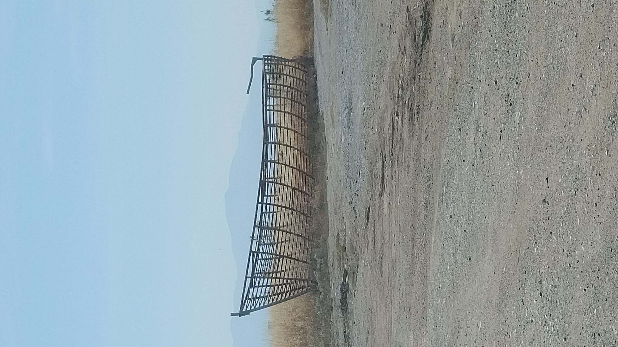 The scene presents a large, skeletal boat-like structure, constructed from dark-colored, horizontally arranged slats forming an open hull, positioned on dry, arid terrain in Thermi, Greece. The structure, located in the midground, has its bow oriented towards the left of the frame and its stern towards the right, where an elevated, curved element is visible on the starboard side. The ground immediately surrounding the structure consists of sparse, dry, light brown vegetation. The foreground is composed of a light gray gravel and dirt path, exhibiting visible granular texture and small stones. In the background, hazy, distant mountains are discernible beneath a vast, pale blue, cloudless sky that occupies the upper half of the image. No individuals or actions are depicted in the scene.