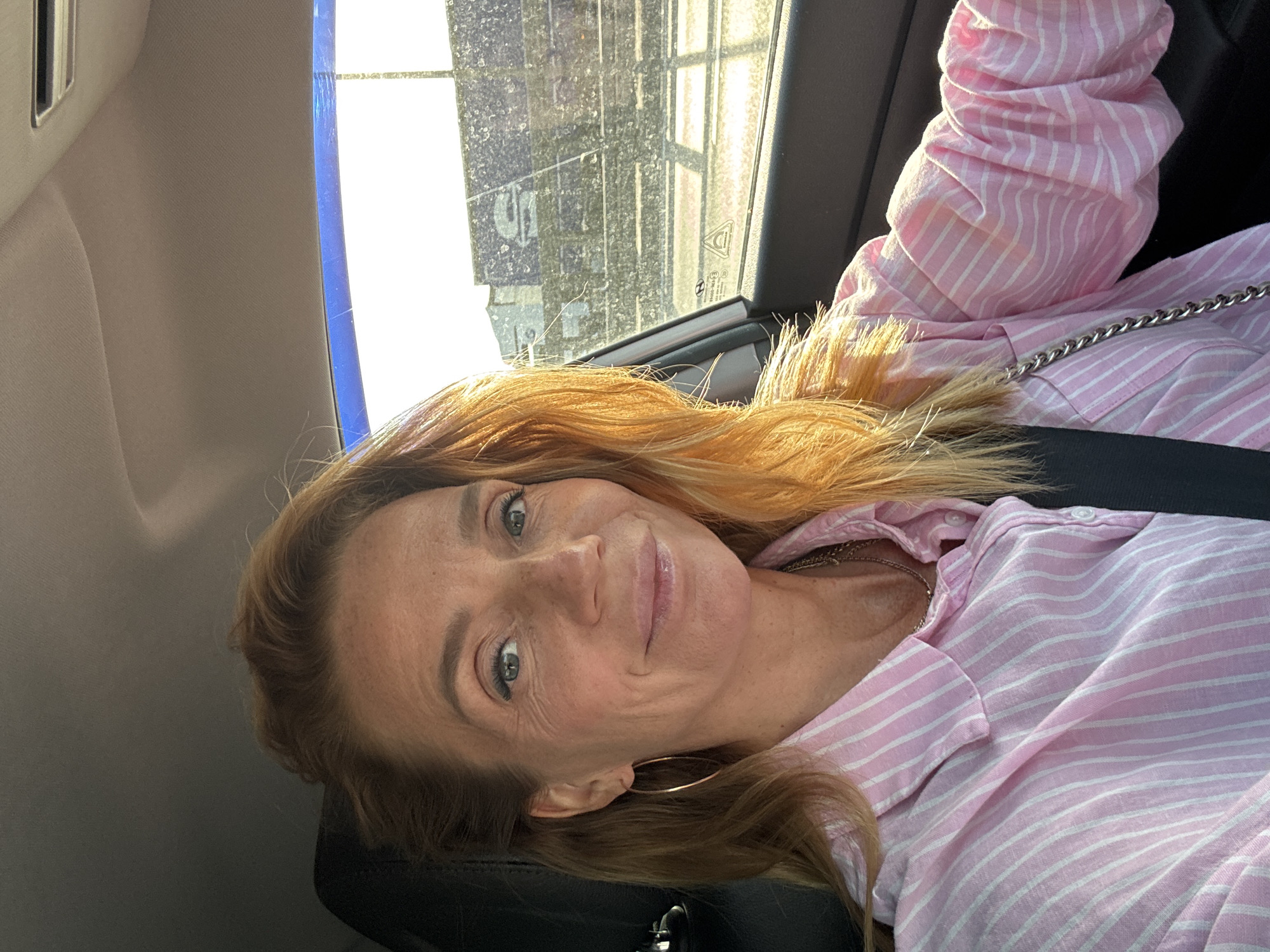 A woman with blonde hair is sitting in the driver's seat of a car, taking a selfie. She is wearing a pink and white striped shirt and a necklace. The sun is shining, creating a bright glare on the right side of the image, and a building is visible in the background through the car window. The car appears to be on a road, possibly in Saint Petersburg, Russia, given the context provided.