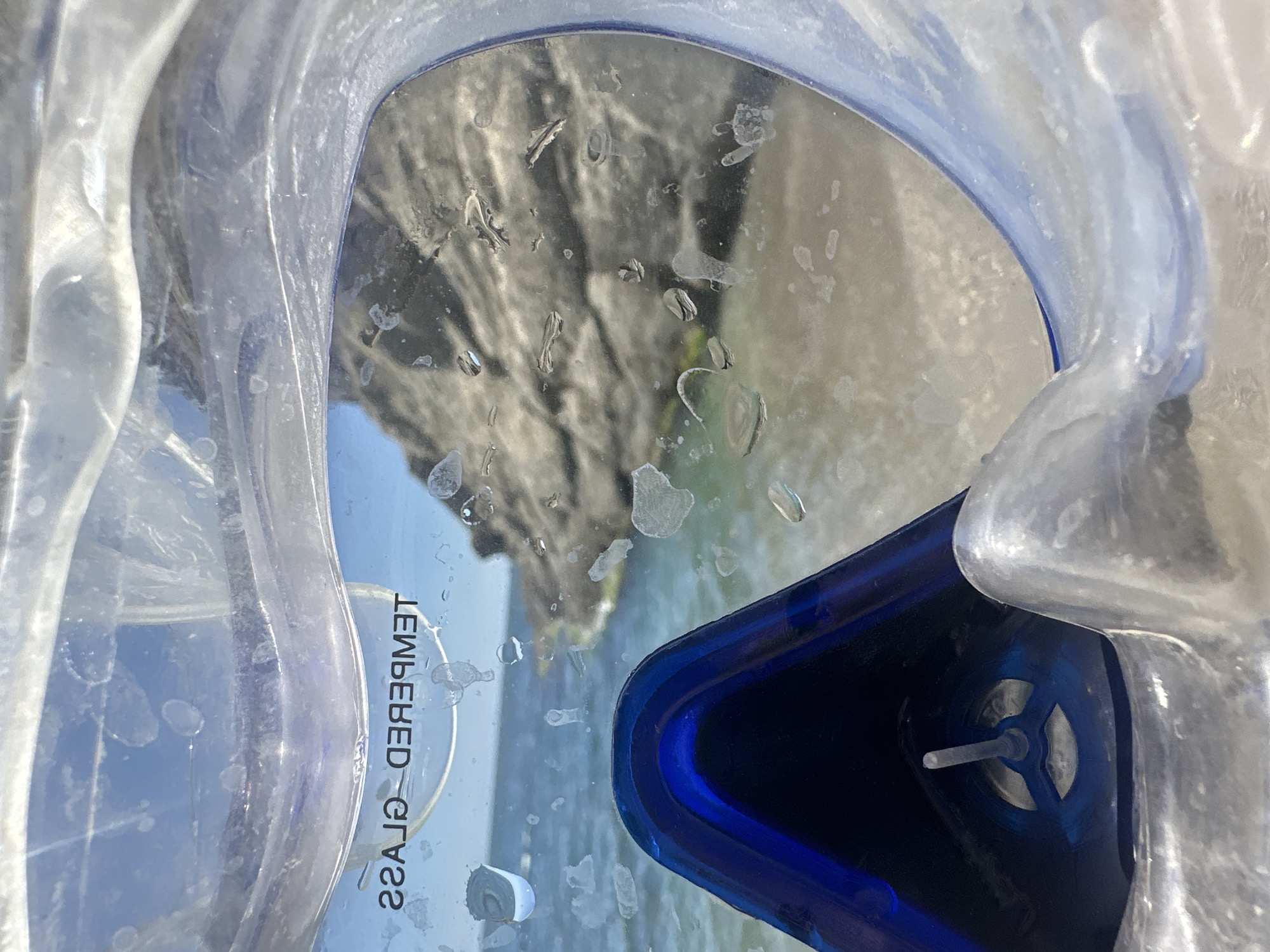 A close-up, interior view of a blue and clear diving mask shows a rocky, coastal landscape through its lens. The mask's silicone skirt and frame are visible, with the blue portion forming a nose pocket and part of the strap attachment. The clear lens has numerous water droplets on its surface, creating distortions and obscuring parts of the scene behind it. In the background, a calm ocean meets a sandy shore, with a large, rugged rock formation jutting out into the water under a clear blue sky. Text on the lens, reading "TEMPERED GLASS" in reverse, is visible. The overall impression is one of a serene beach scene viewed from the perspective of someone about to enter the water.