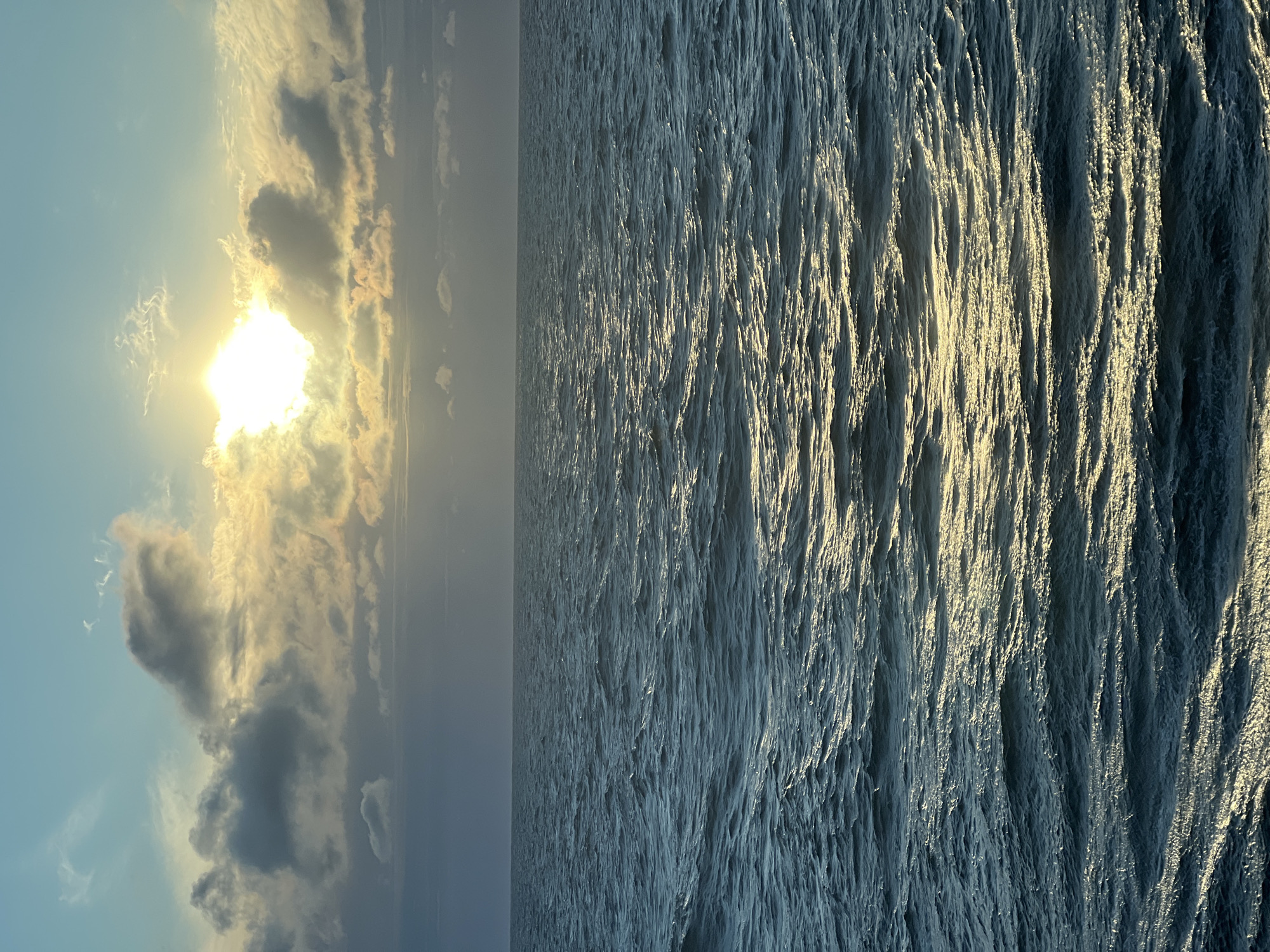 The image depicts a vast, choppy ocean under a sky illuminated by a bright sun partially obscured by clouds. The sun's rays cast a golden sheen across the water's surface, highlighting the texture of the waves. The sky is a gradient of light blue and grey, with dramatic cloud formations catching the sunlight. The horizon is a sharp, clear line separating the sea from the sky. There are no people, structures, or landmasses visible in the image. The scene conveys a sense of natural beauty and the immensity of the ocean. The location is stated to be Unknown, Poland, but the image itself provides no specific geographical markers to confirm this.