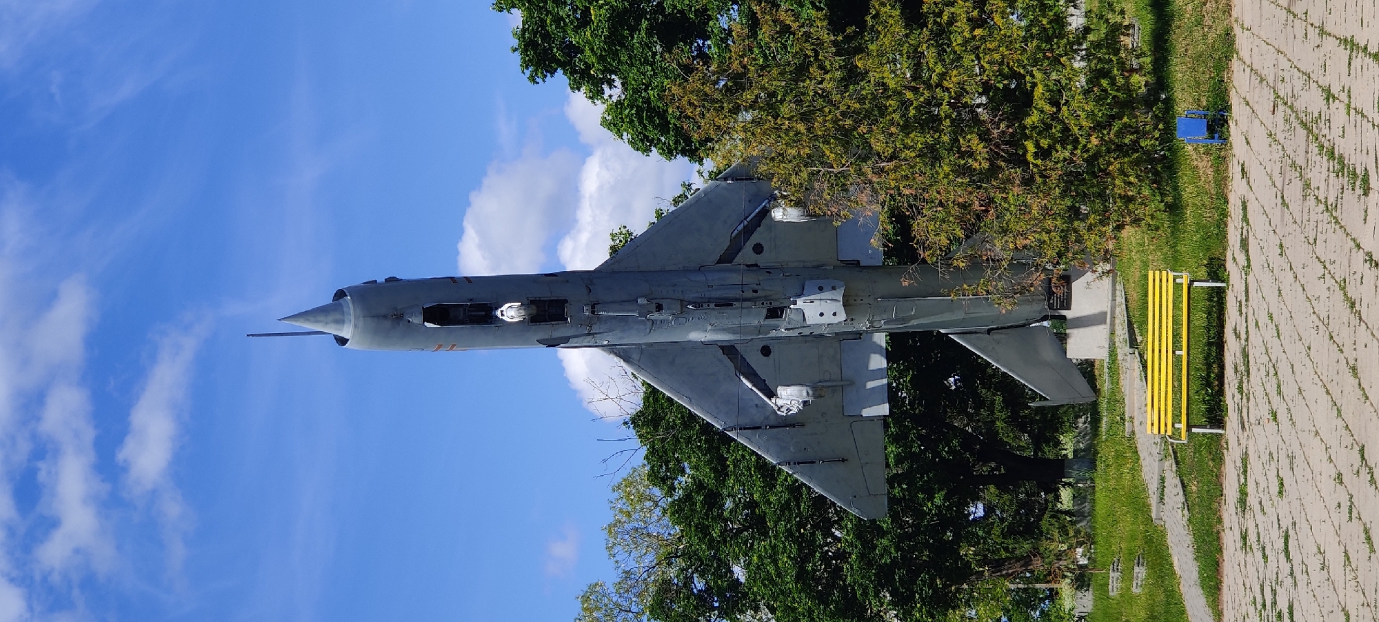 A MiG-21 fighter jet is displayed on a concrete plinth in Kolomak, Ukraine. The aircraft is painted in gray camouflage. A yellow bench is located nearby. Lush trees surround the aircraft, partially obscuring it from view. A blue sky with white clouds forms the backdrop. A paved walkway is visible to the right.