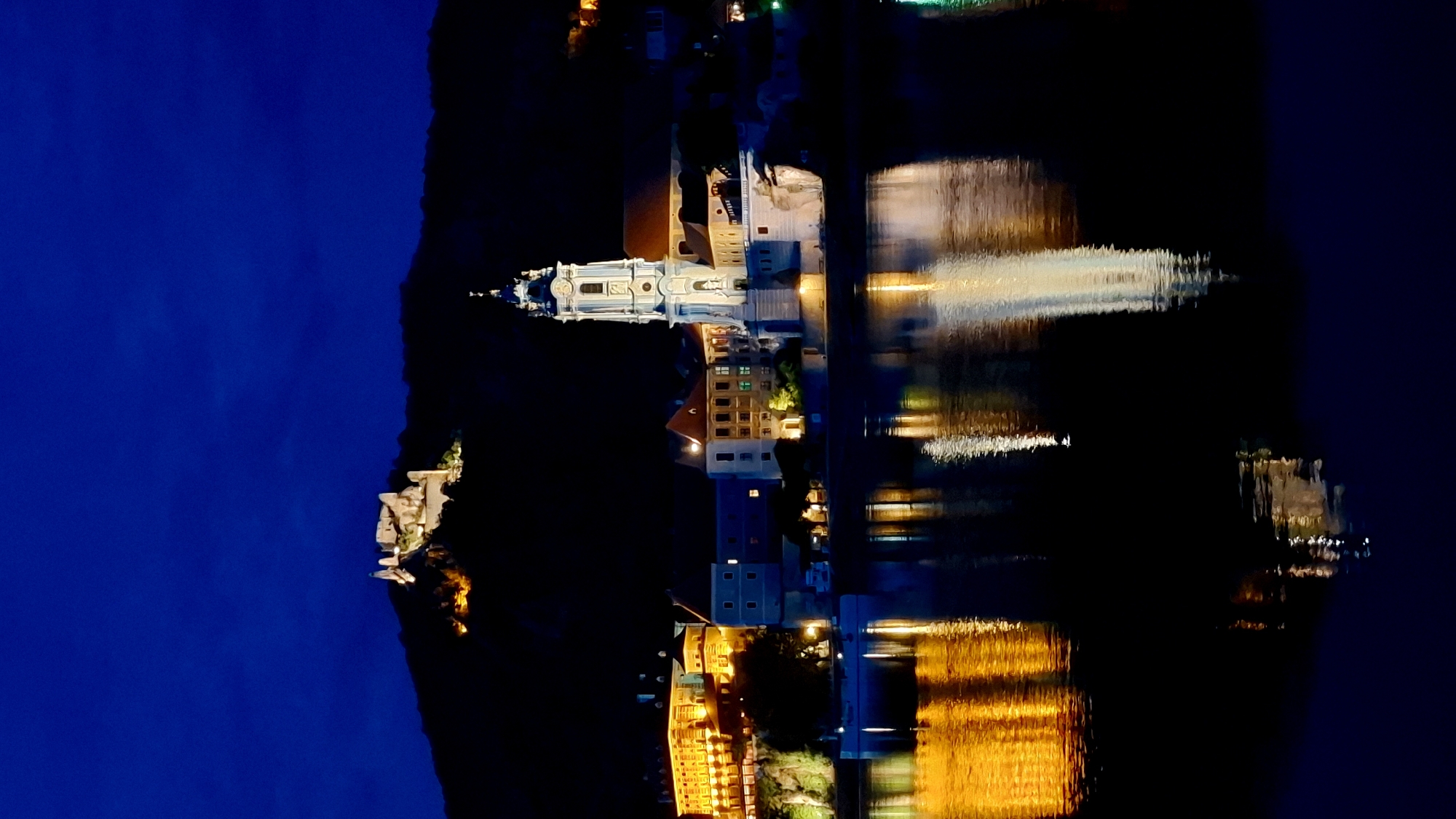 The image is a nighttime view of Rossatz-Arnsdorf, Austria, showcasing the Wachau Valley and the Danube River. In the background, a historic castle ruin is illuminated on a hill. In the midground, a prominent church with a tall, white bell tower stands on the riverbank, flanked by other illuminated buildings. The dark sky is a deep blue, with some faint clouds. The foreground features the dark, reflective surface of the Danube River, mirroring the lights of the town and the church tower. The reflections create streaks of yellow, white, and orange on the water. There are no people visible in the image. The scene depicts a tranquil and picturesque European riverside town at night, with historical landmarks prominently featured.